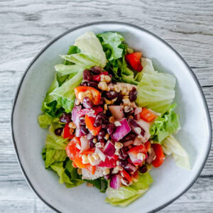 A close-up of a cowboy caviar chicken salad featuring chopped lettuce topped with a colorful mix of black beans, red onions, red bell peppers, corn, and diced chicken, all served in a white bowl on a wood-grain table.