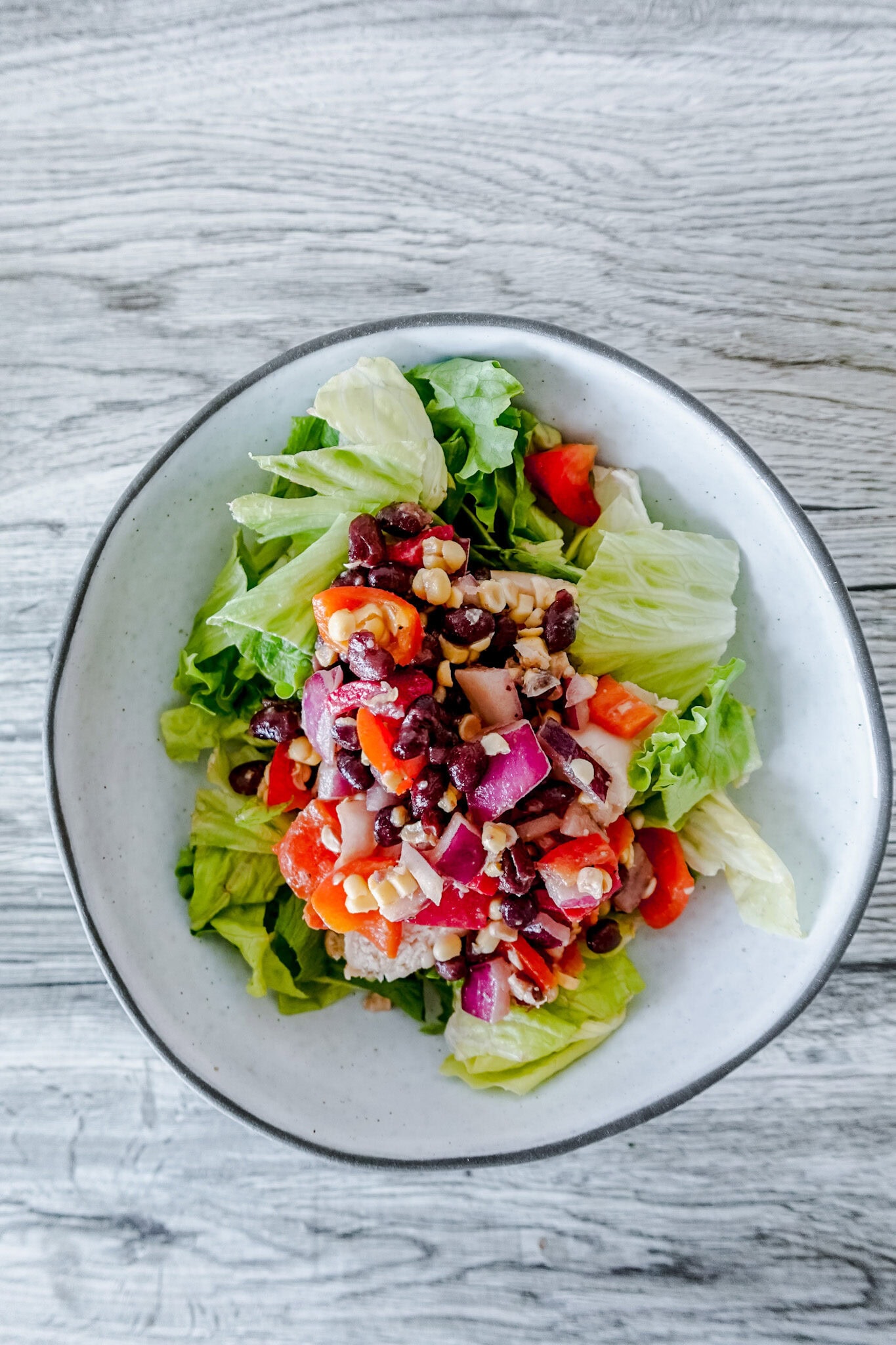 A close-up of a cowboy caviar chicken salad featuring chopped lettuce topped with a colorful mix of black beans, red onions, red bell peppers, corn, and diced chicken, all served in a white bowl on a wood-grain table.