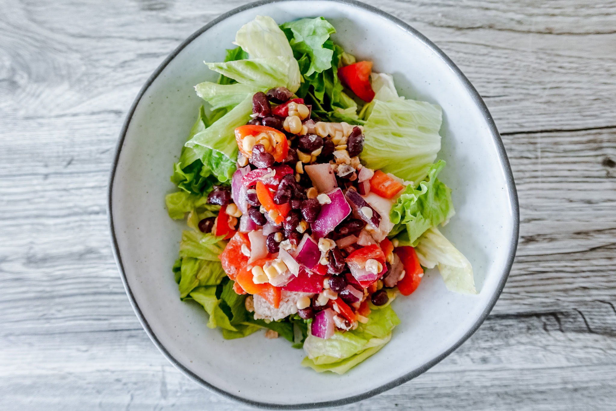 A vertical view of cowboy caviar chicken salad, showcasing layers of crisp lettuce, chunks of chicken, and a vibrant topping of black beans, red peppers, onions, and corn in a shallow white bowl.