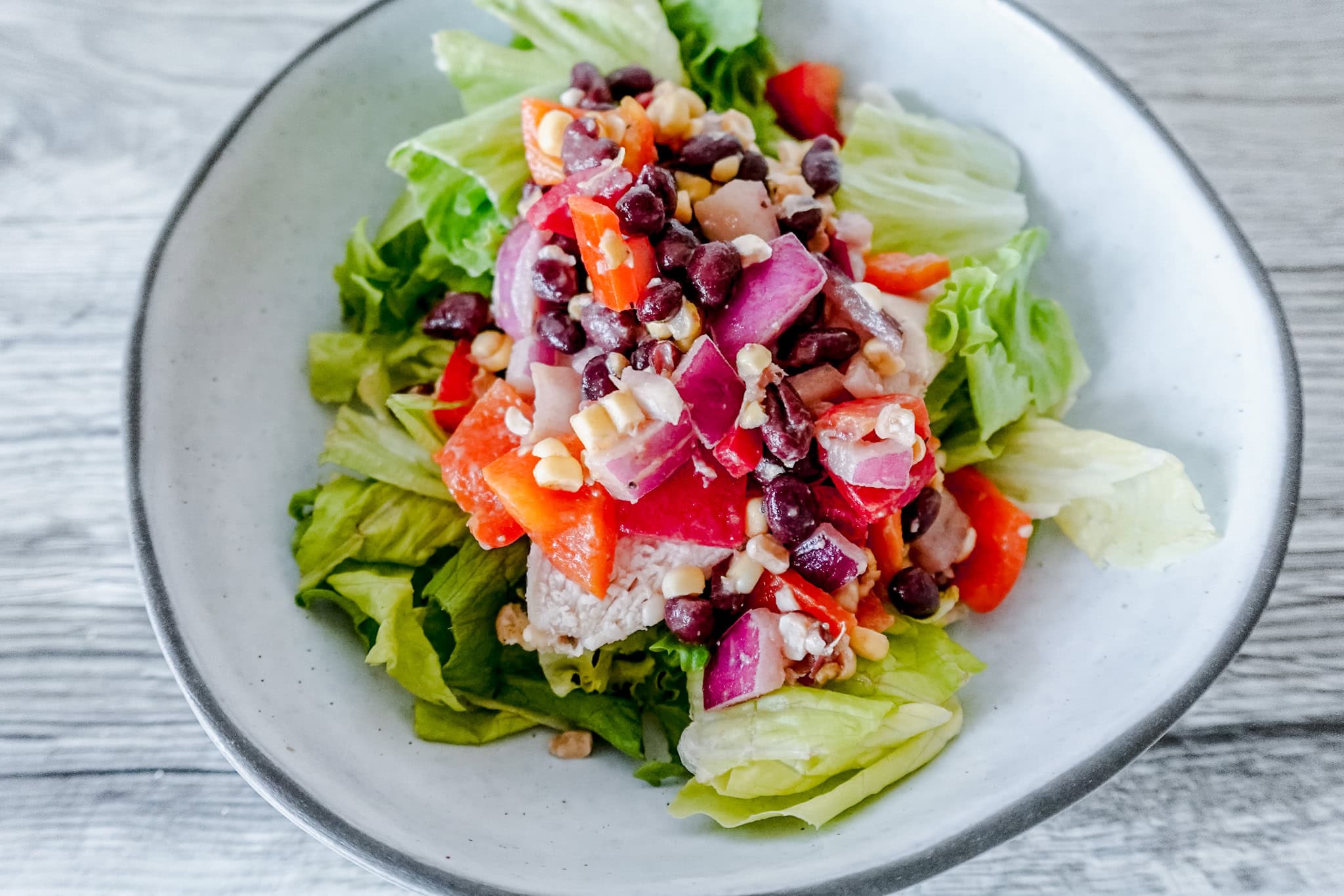 An angled close-up shot of cowboy caviar chicken salad with hearty pieces of chicken, black beans, red onion, corn, and bell pepper arranged on a bed of green lettuce in a white dish.