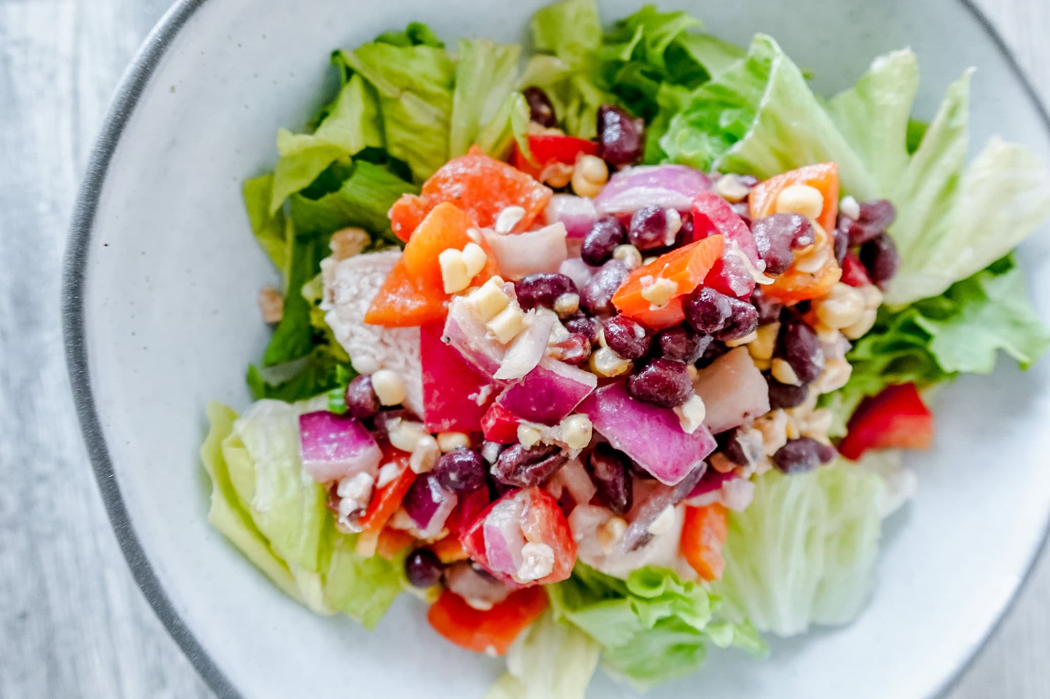 A bright, top-down view of cowboy caviar chicken salad, with the ingredients neatly layered in a white bowl: crisp lettuce, chunks of chicken, and a colorful cowboy caviar topping.