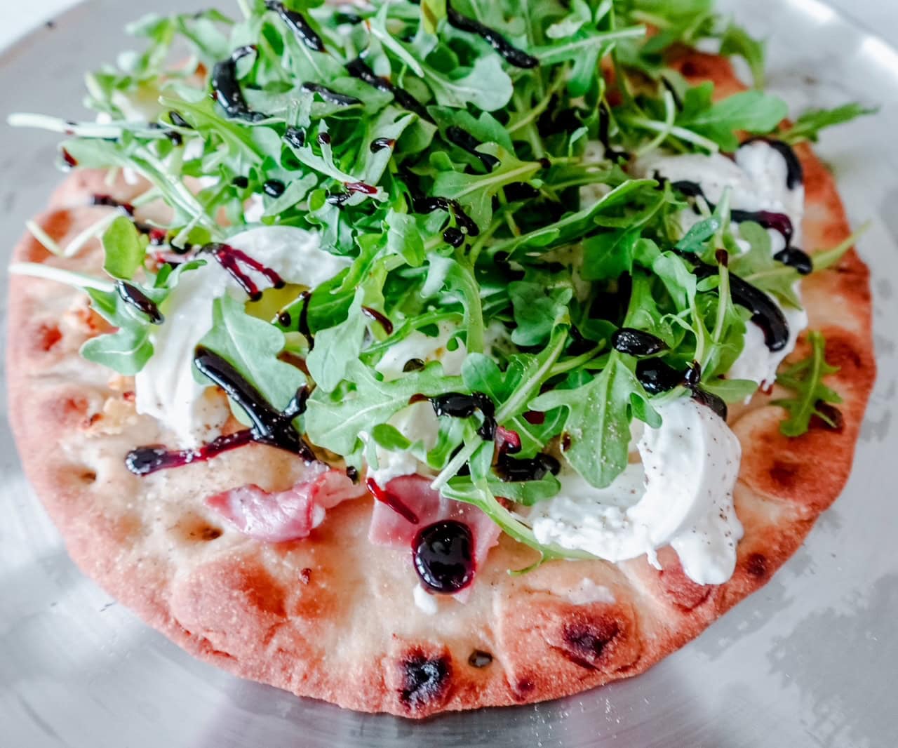 Overhead view of a rustic burrata and prosciutto pizza on a metal tray, topped generously with fresh arugula and drizzled with balsamic glaze.