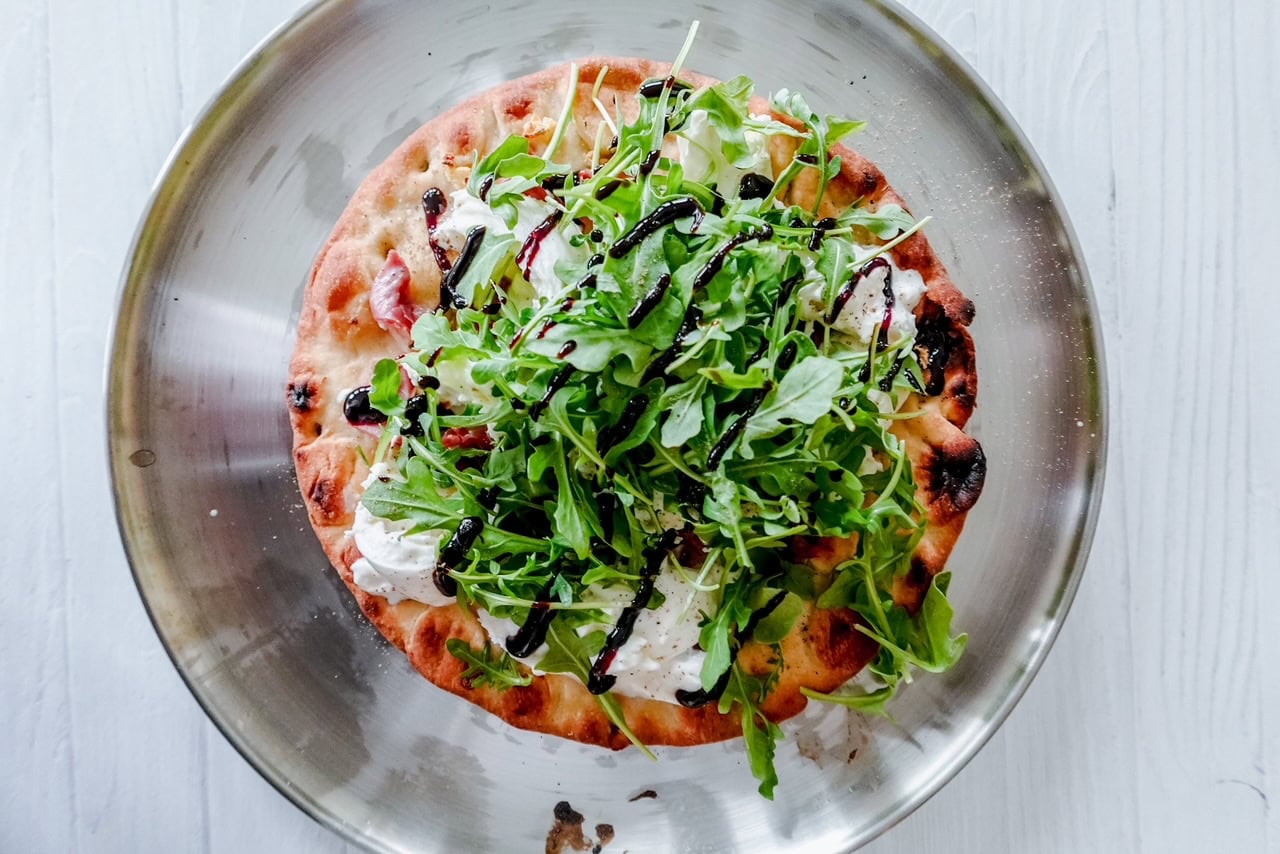 Tight zoom on folded prosciutto nestled among arugula and creamy burrata on a charred, blistered flatbread crust with balsamic drizzle.