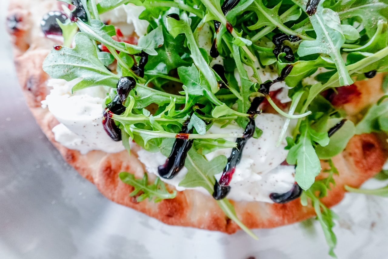 Side angle of burrata and prosciutto pizza showcasing the airy flatbread crust, thick layers of burrata, and bright green arugula with a dark balsamic drizzle.