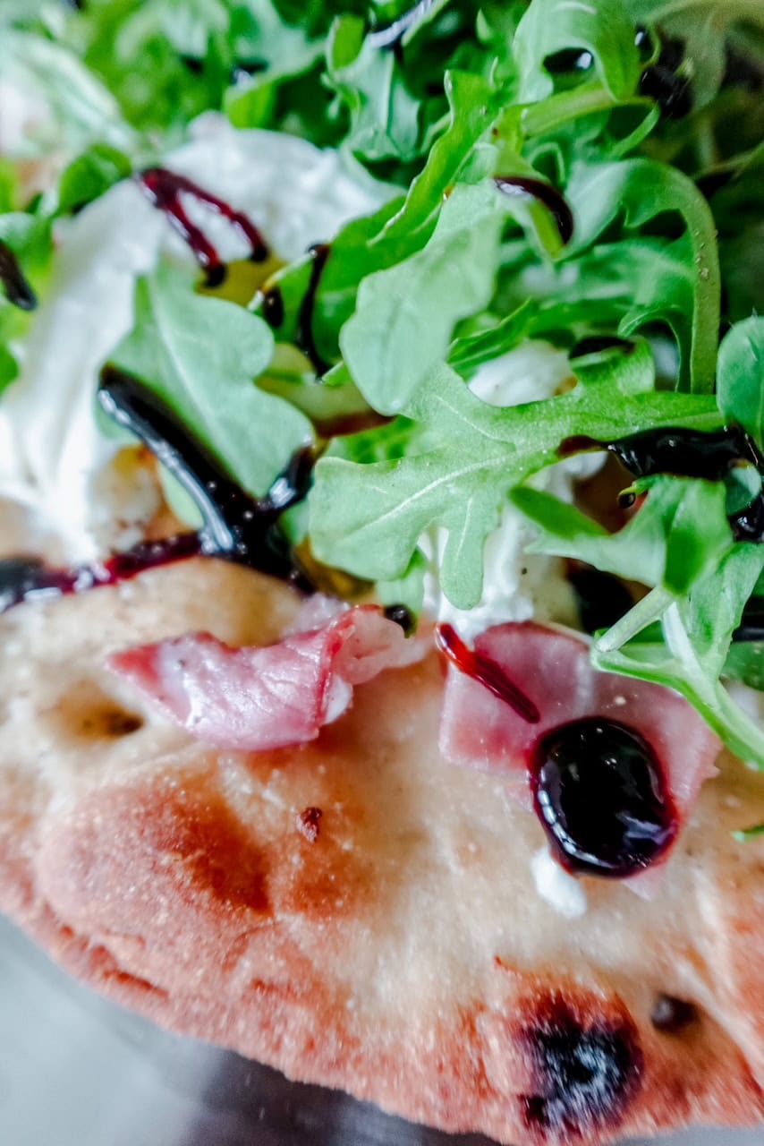 Macro image capturing texture details of creamy burrata, peppery arugula, and sticky balsamic glaze on crispy flatbread with bits of prosciutto peeking through.