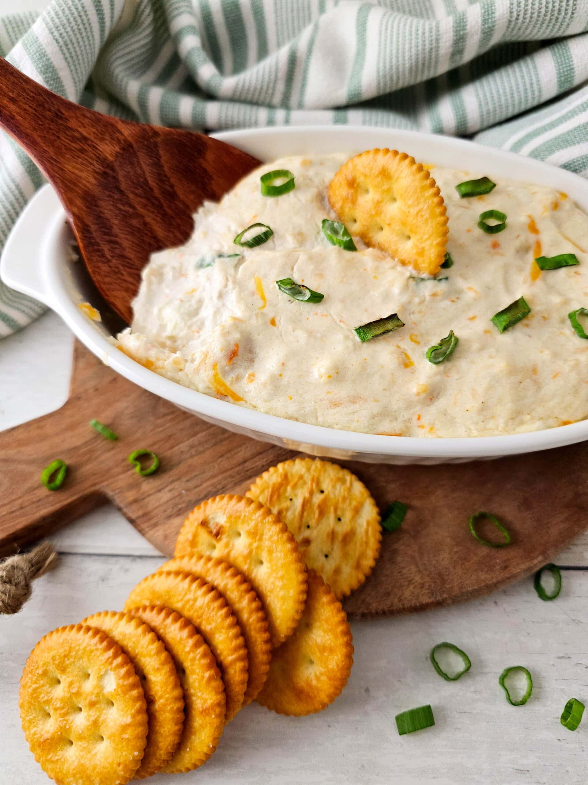 lose-up of a creamy hot seafood dip garnished with chopped green onions in a white baking dish, with a golden round cracker partially dipped inside and a wooden spoon resting in the dip.