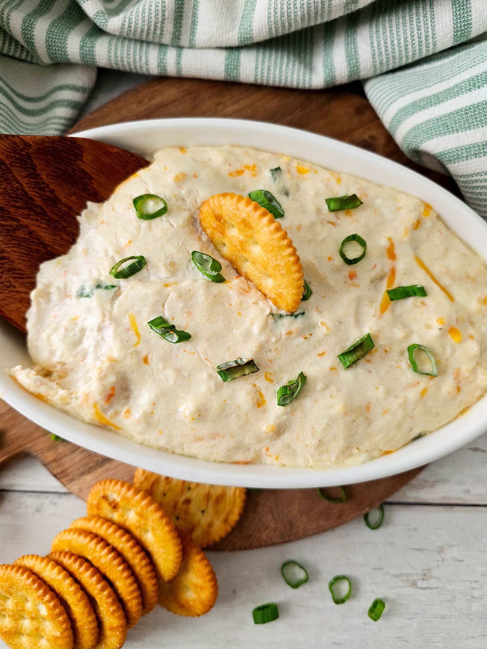 Overhead view of hot seafood dip in a white oval dish, topped with sliced green onions and a cracker, surrounded by stacked crackers and set on a wooden board with a green and white striped kitchen towel in the background.