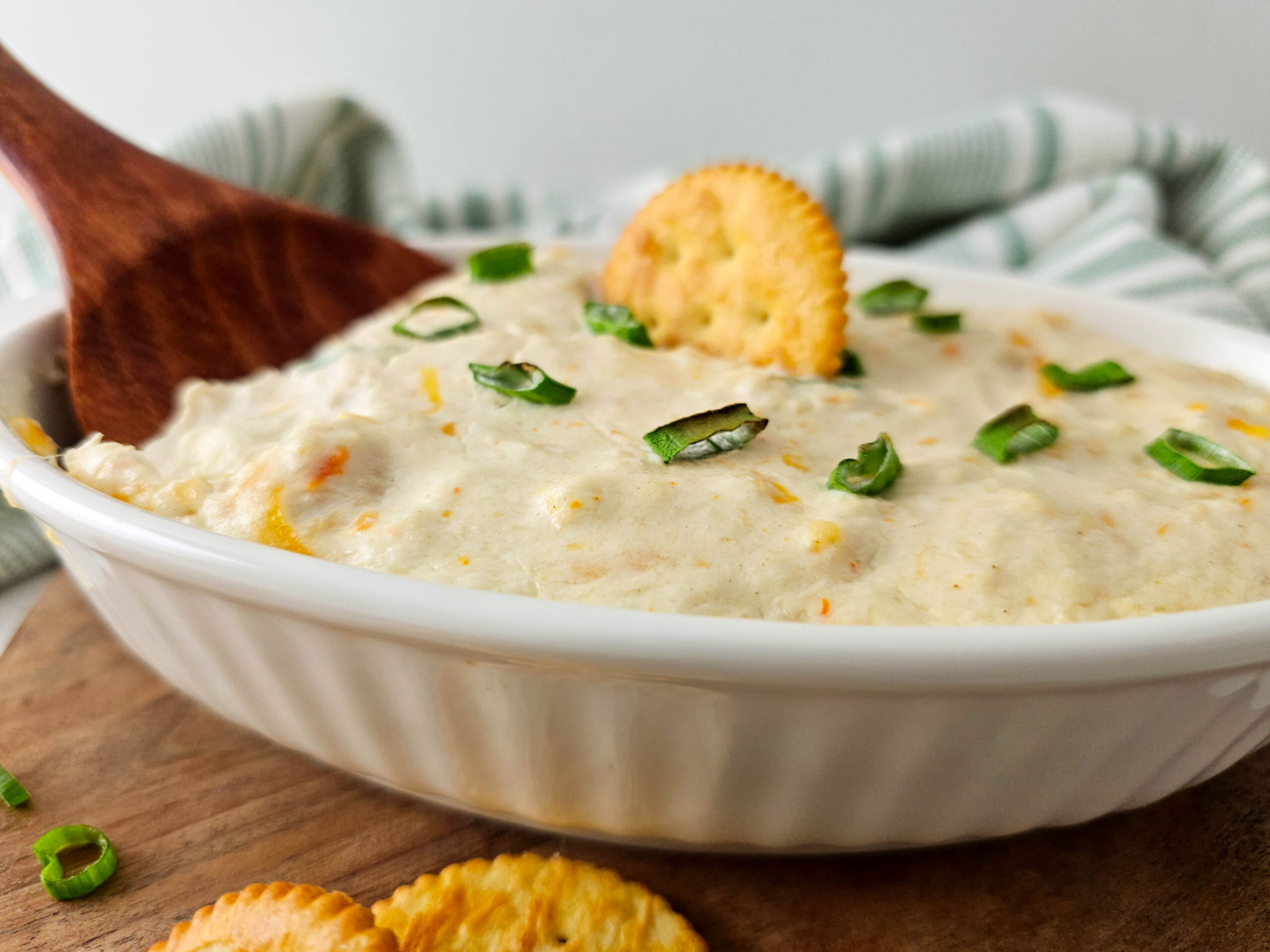 Top-down view of the hot seafood dip with a cracker placed in the center and sliced green onions on top, accompanied by a stack of round crackers on a wooden board.