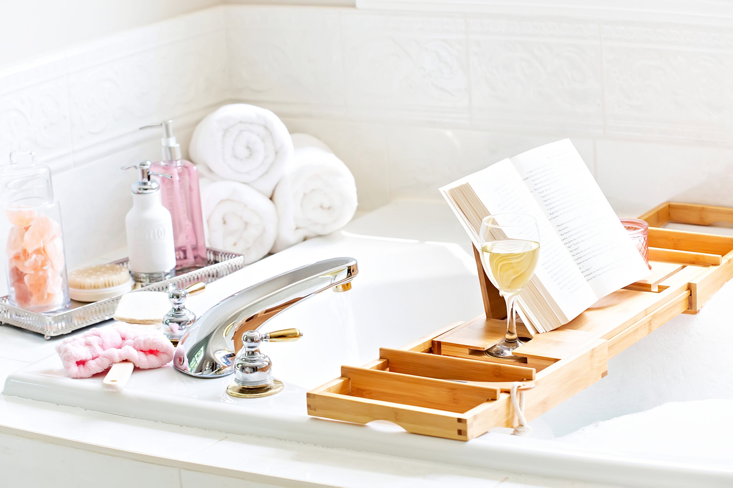 Luxurious bathtub scene with a bamboo bath tray holding a book and a glass of white wine, surrounded by rolled white towels, spa brushes, a glass jar of pink Himalayan salt, and stylish soap dispensers.