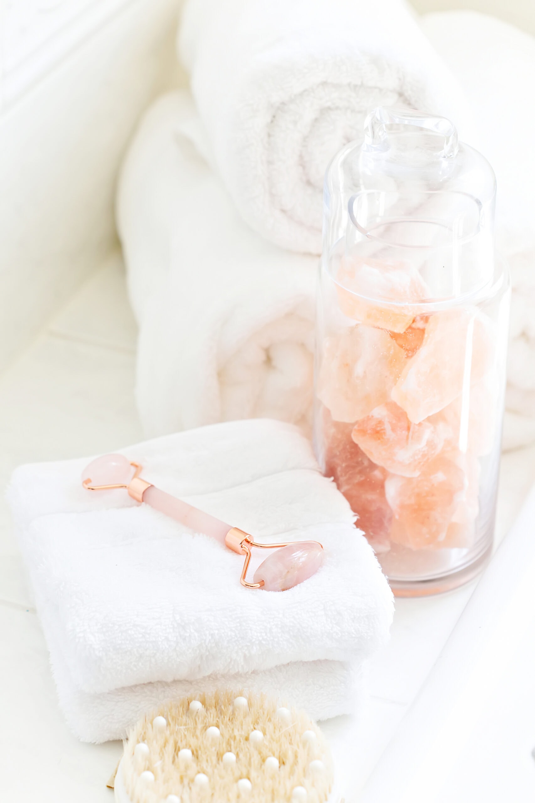 Soft white towels rolled neatly next to a glass container of Himalayan pink salt and a rose quartz face roller resting on stacked towels, creating a serene and indulgent spa vibe.