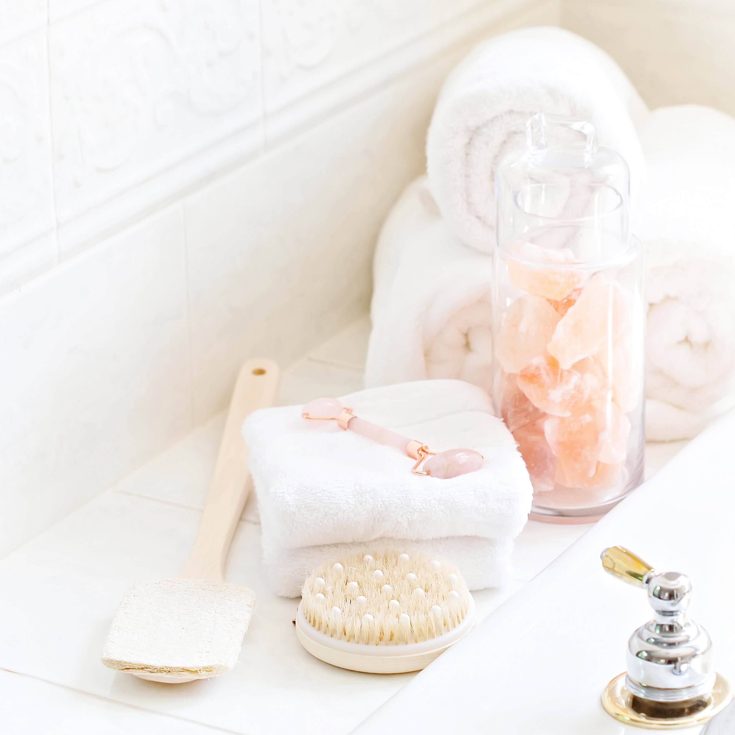 Elegant spa setup featuring white rolled towels, a dry brush, a rose quartz face roller on folded white towels, and a clear jar filled with Himalayan pink salt chunks, all arranged on a clean tiled surface.