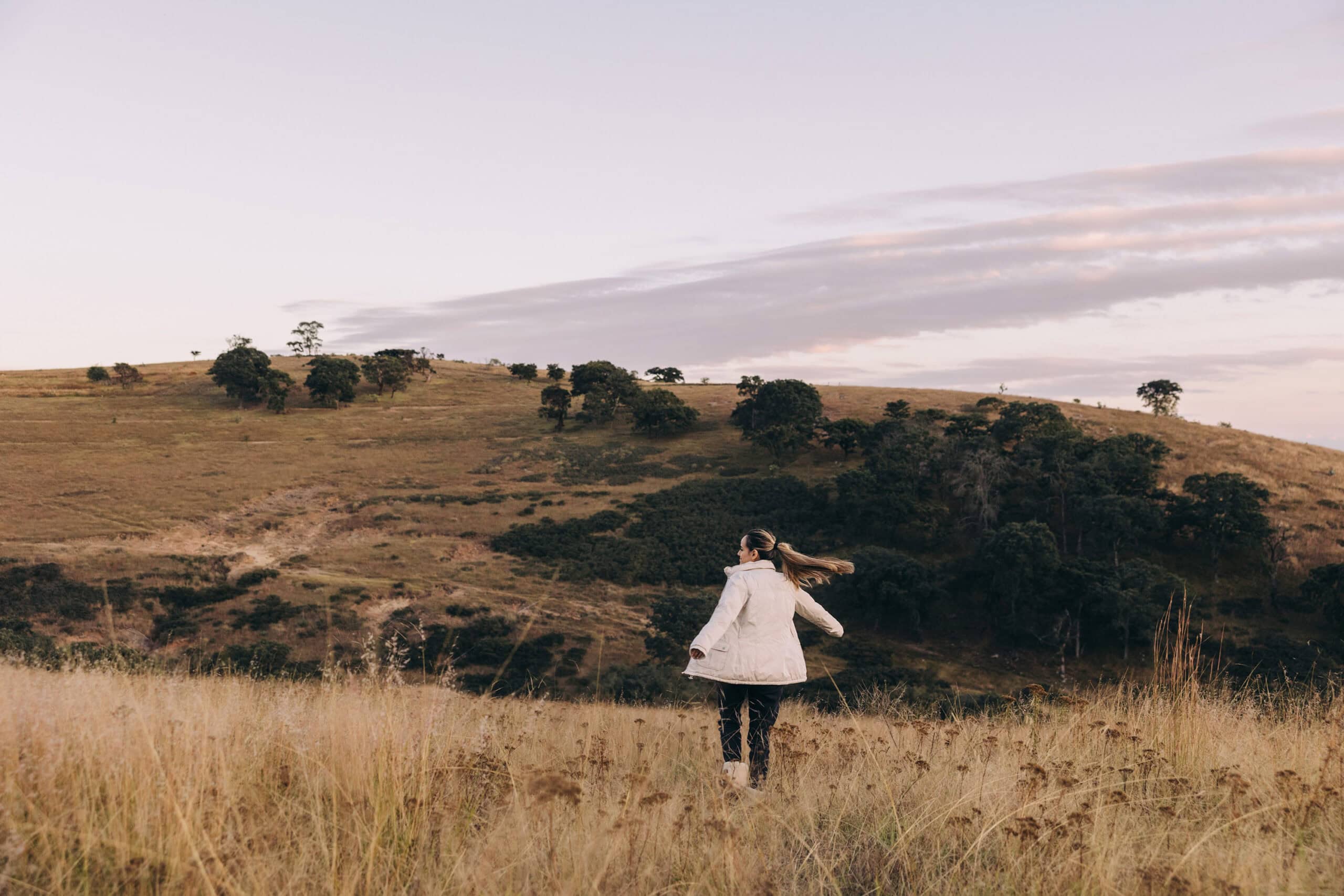 A woman in a white coat walking through a golden, grassy field with rolling hills and scattered trees in the distance under a pastel sky.