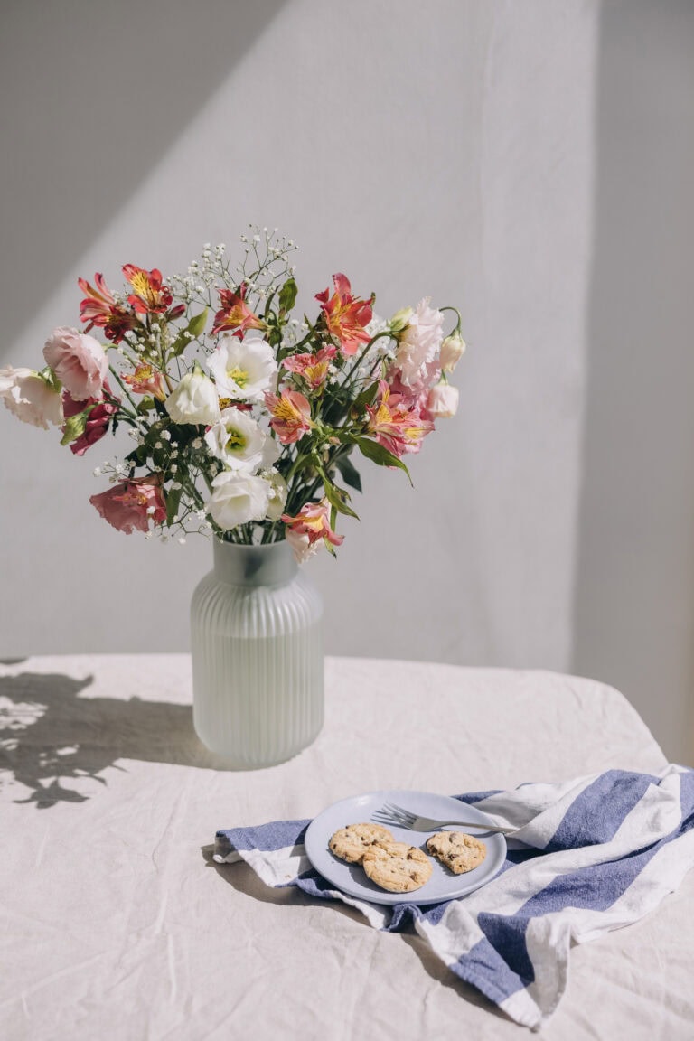 A bouquet of pink, white, and red flowers in a ribbed ceramic vase on a sunlit linen-covered table, with a blue plate of chocolate chip cookies and a striped napkin beside it.