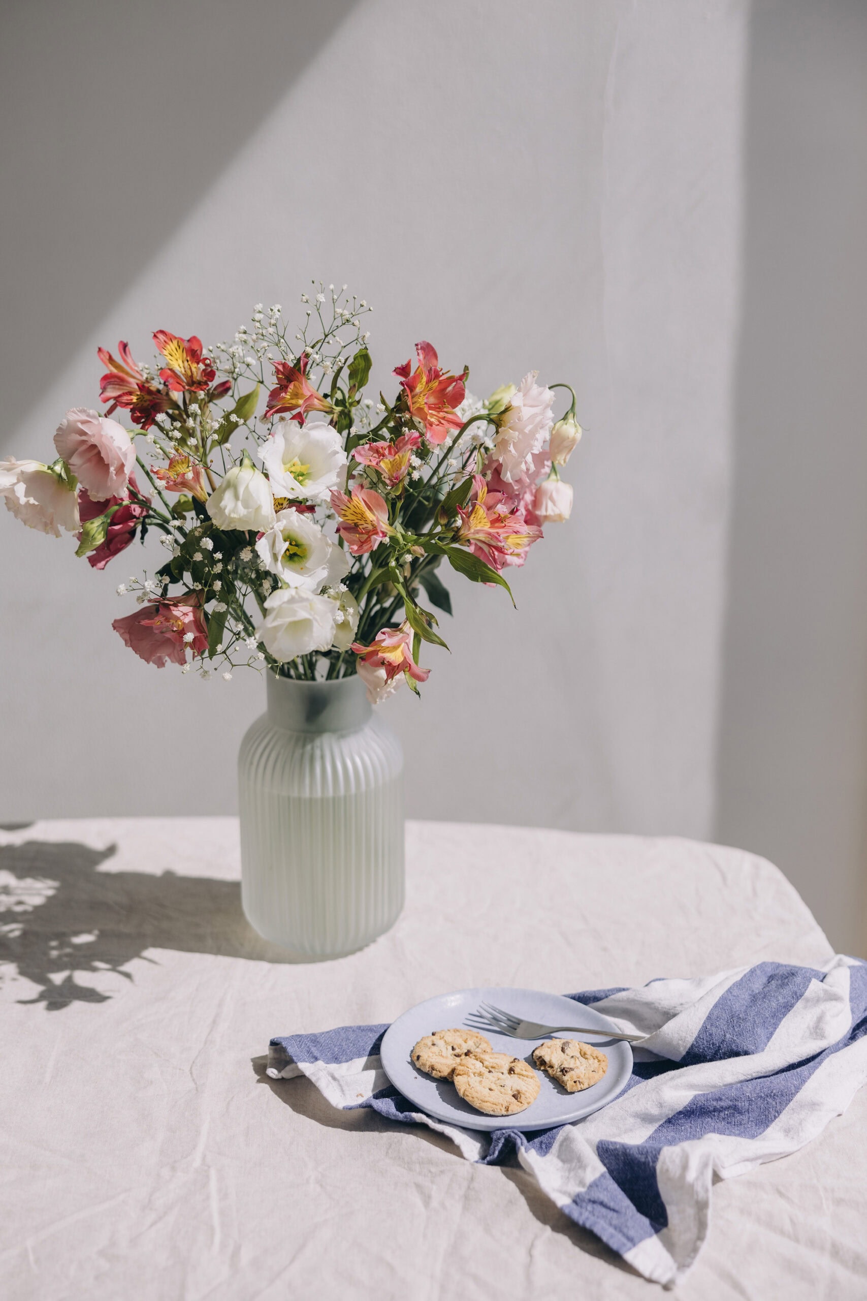 A bouquet of pink, white, and red flowers in a ribbed ceramic vase on a sunlit linen-covered table, with a blue plate of chocolate chip cookies and a striped napkin beside it.