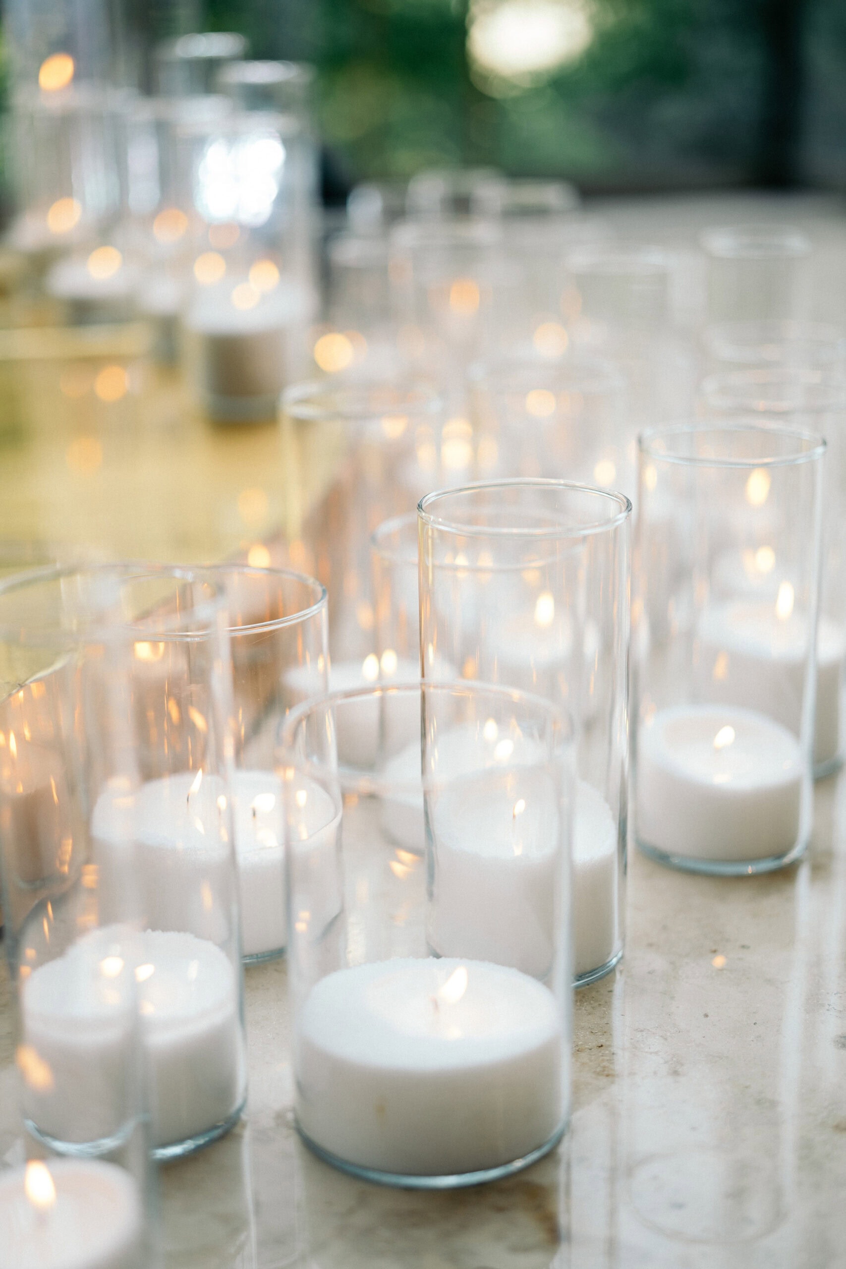 Multiple white votive candles softly glowing inside tall, clear glass holders arranged on a reflective surface, creating a serene and romantic ambiance.