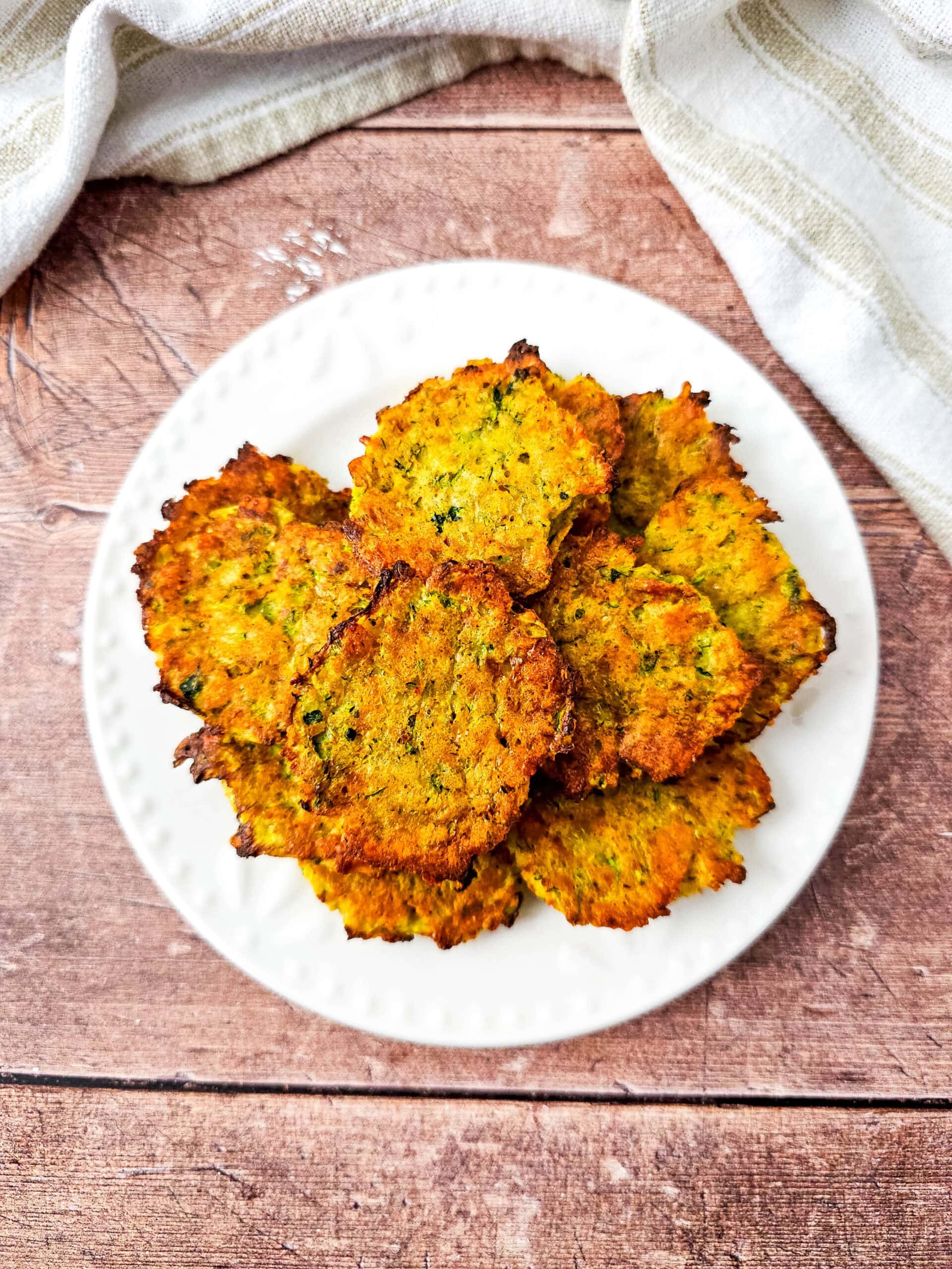 Top-down view of a plate of zucchini tots, perfectly browned and stacked on a white plate over a wooden table with a cream-striped cloth.