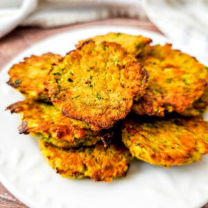 Close-up side view of crispy zucchini tots stacked on a white plate, showing their golden exterior and flecks of green zucchini.