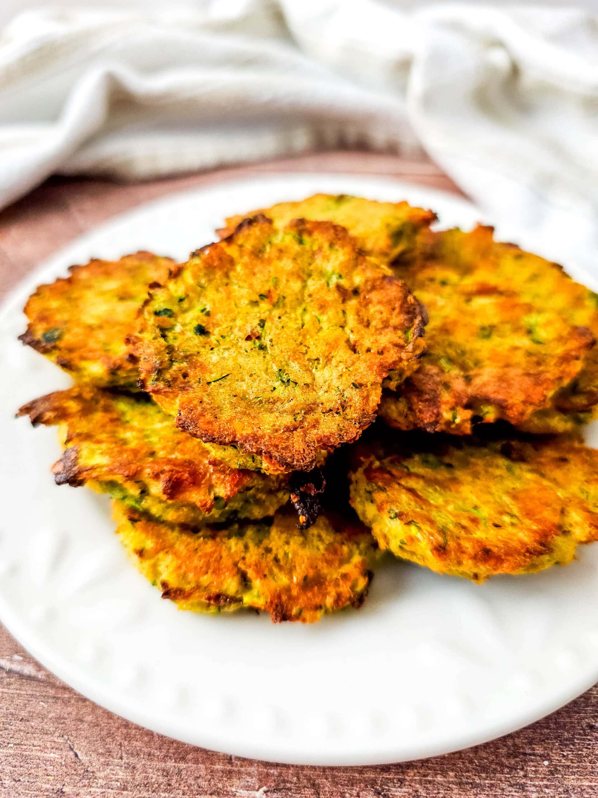 Close-up side view of crispy zucchini tots stacked on a white plate, showing their golden exterior and flecks of green zucchini.