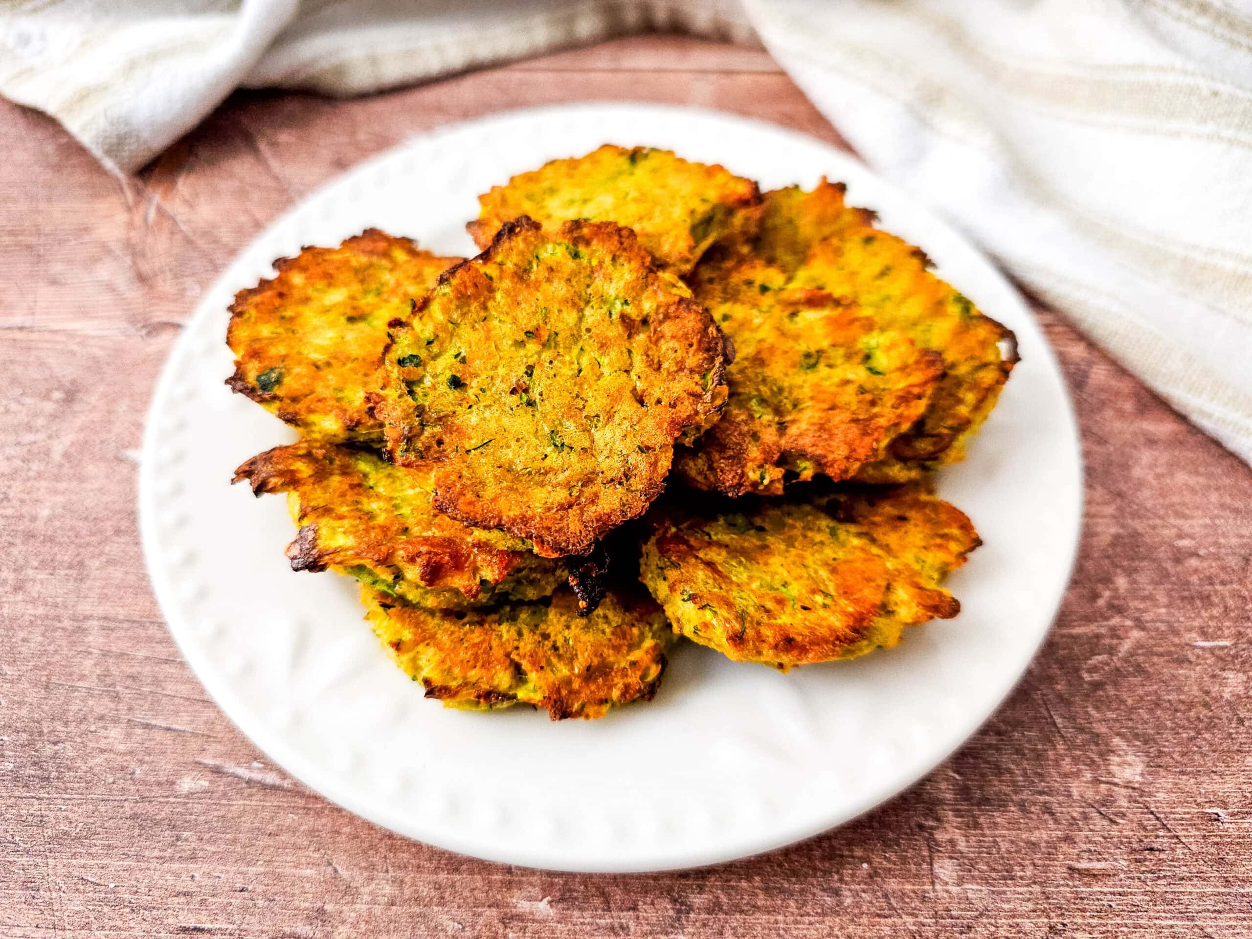 Overhead image of golden zucchini tots arranged in a circular pattern on a white plate, styled on a wooden table with a striped towel.