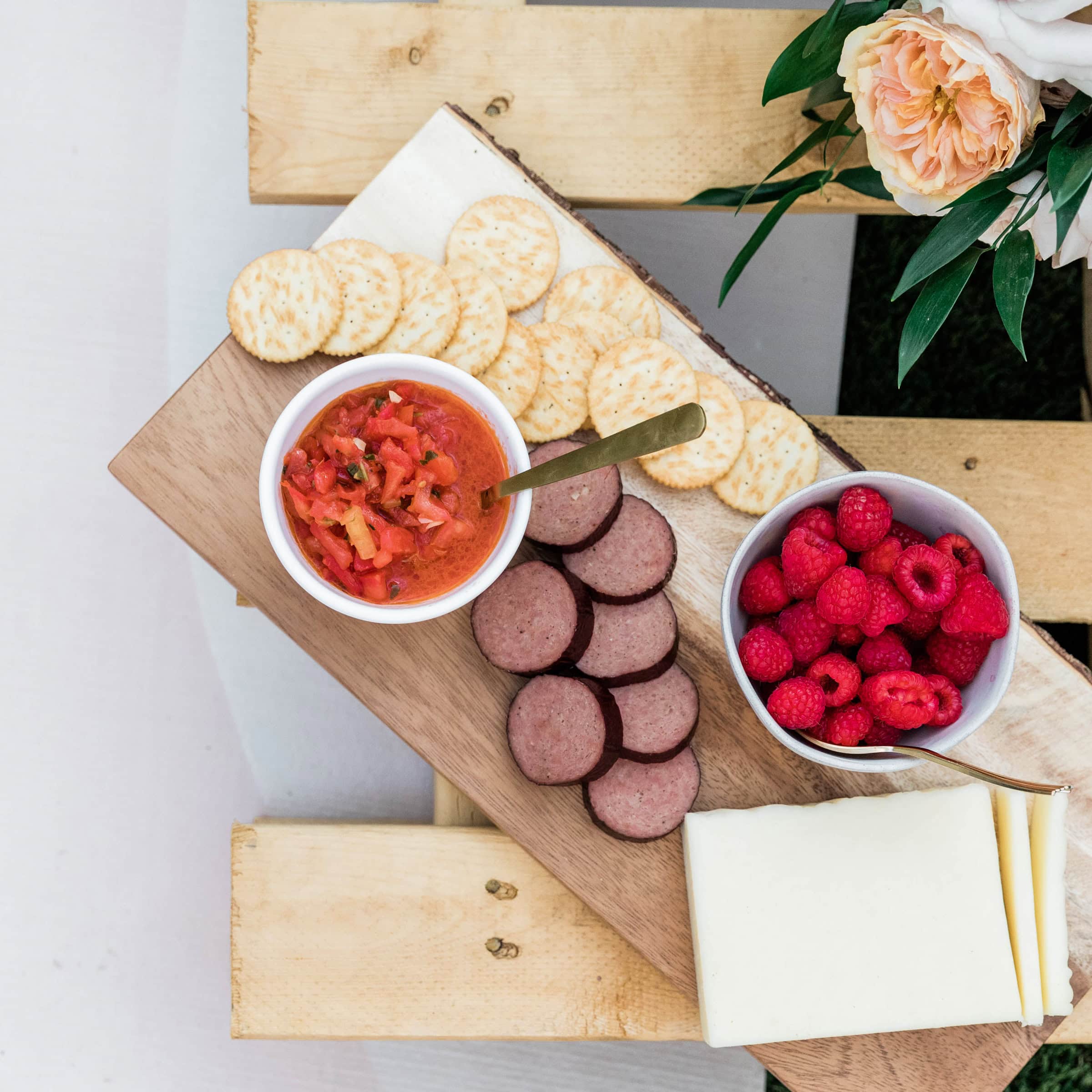 A rustic wooden board displays a selection of crackers, sliced sausage, cheese, fresh raspberries, and a bowl of tomato relish, capturing the relaxed, communal spirit of French outdoor dining.