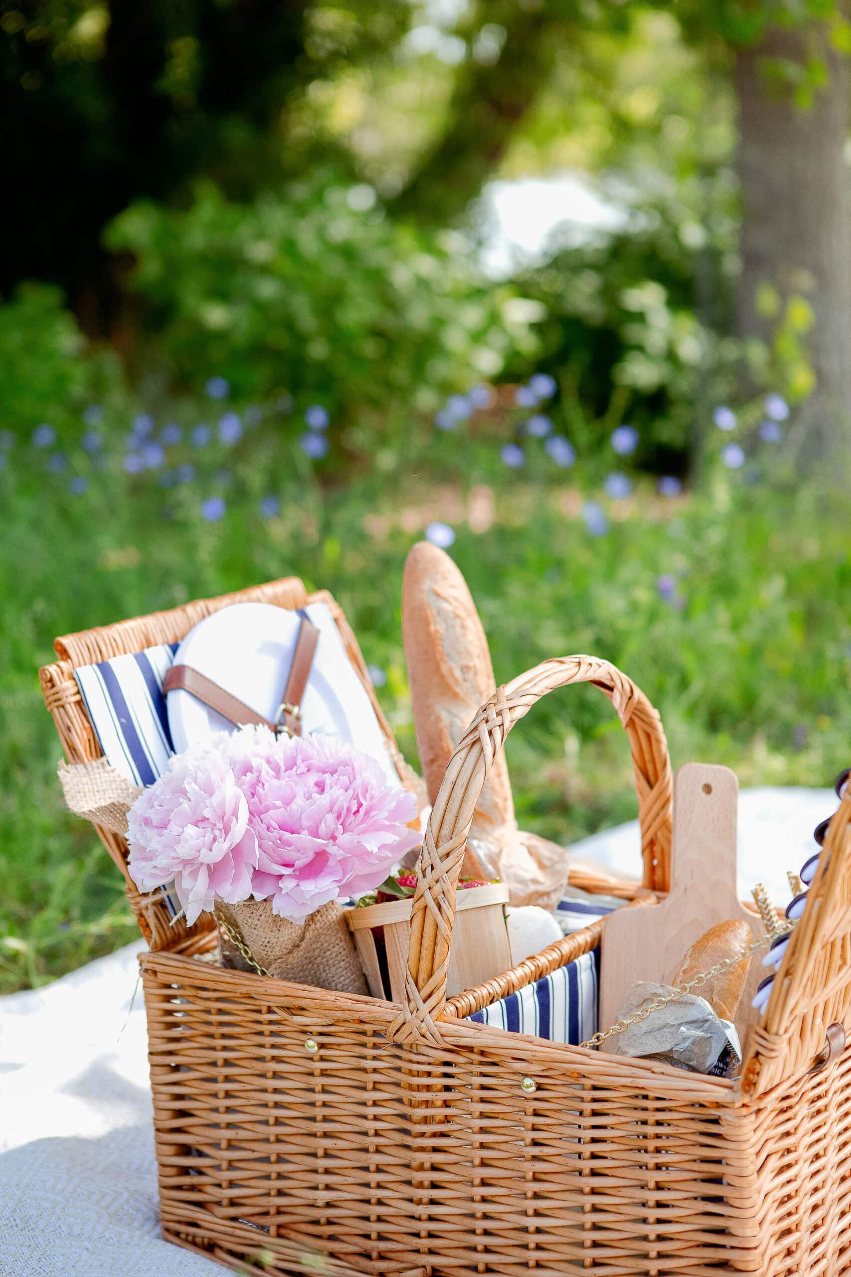 A charming wicker picnic basket sits on a blanket in a sunny meadow, filled with baguettes, striped napkins, and a bouquet of pink peonies—evoking the effortless elegance of a French summer picnic.