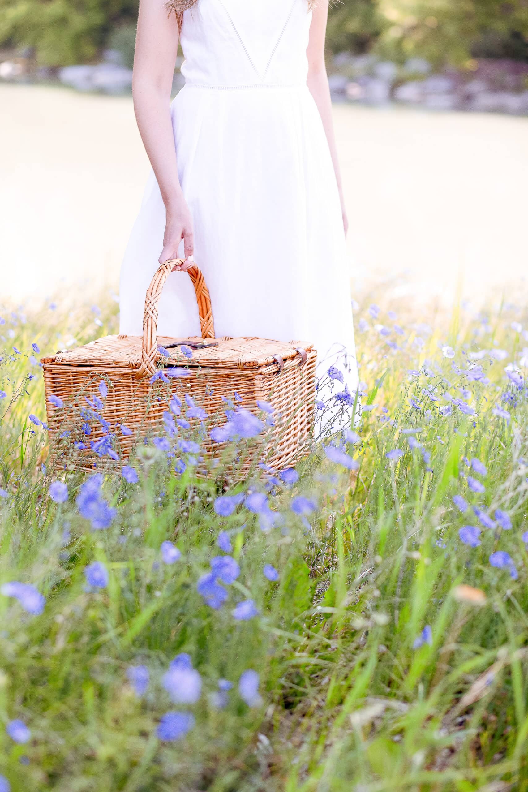 A woman in a white summer dress stands in a sunlit field of wildflowers, holding a woven picnic basket—capturing the essence of relaxed, French-inspired summer living.