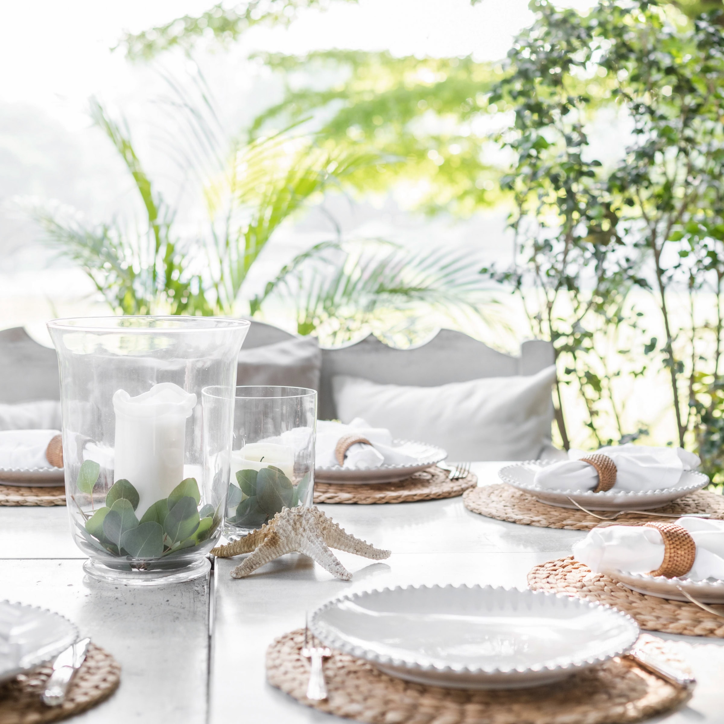A bright, airy outdoor table is set with woven placemats, white plates, and glass hurricane candle holders filled with greenery, creating a simple yet sophisticated French-inspired dining atmosphere.