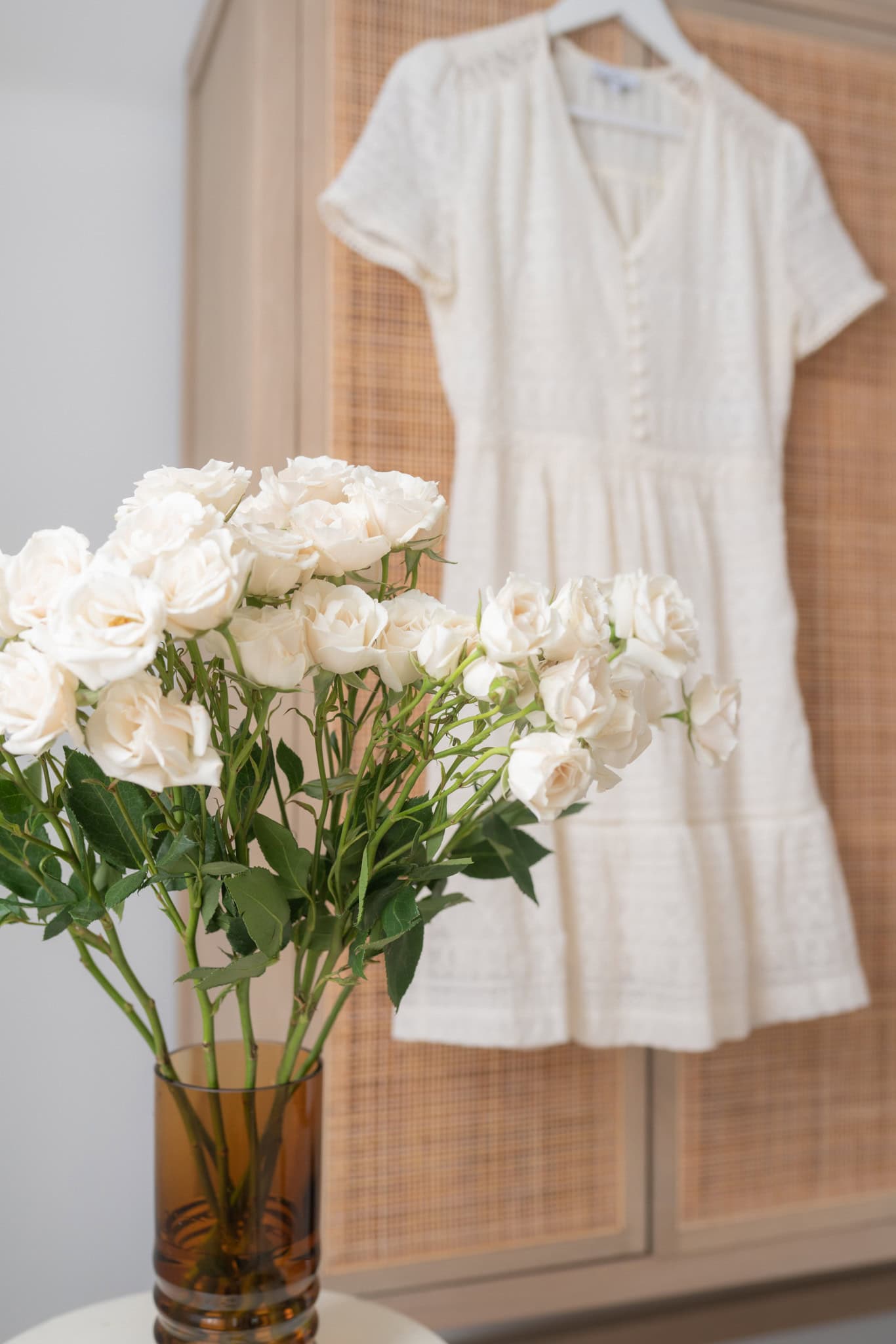 A bouquet of creamy white roses in a brown glass vase is placed in front of a softly blurred white summer dress, hinting at the understated chic and everyday beauty of French living.