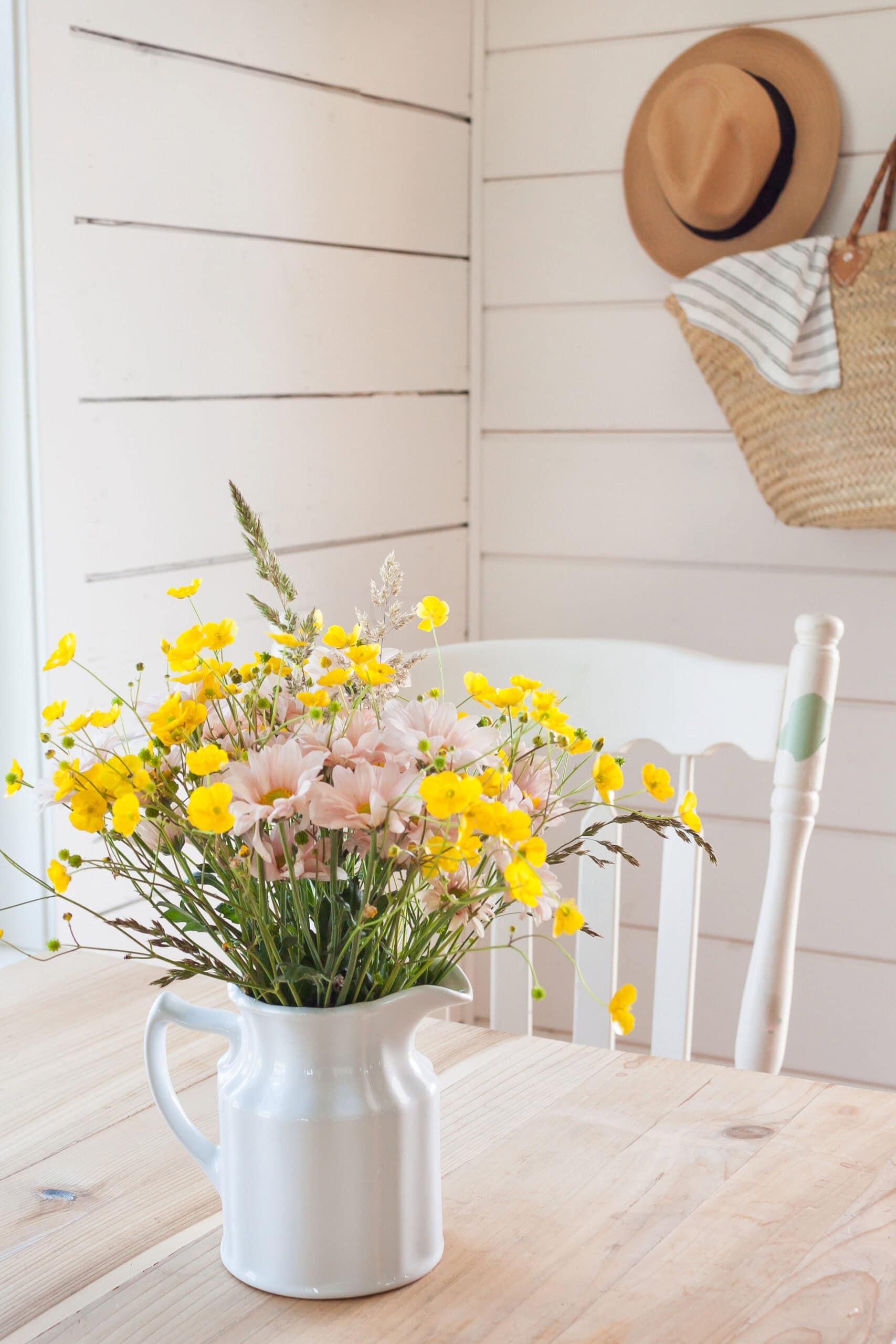 A white ceramic pitcher filled with delicate yellow and blush wildflowers sits on a wooden farmhouse table, channeling the effortless, natural beauty found in French country homes.