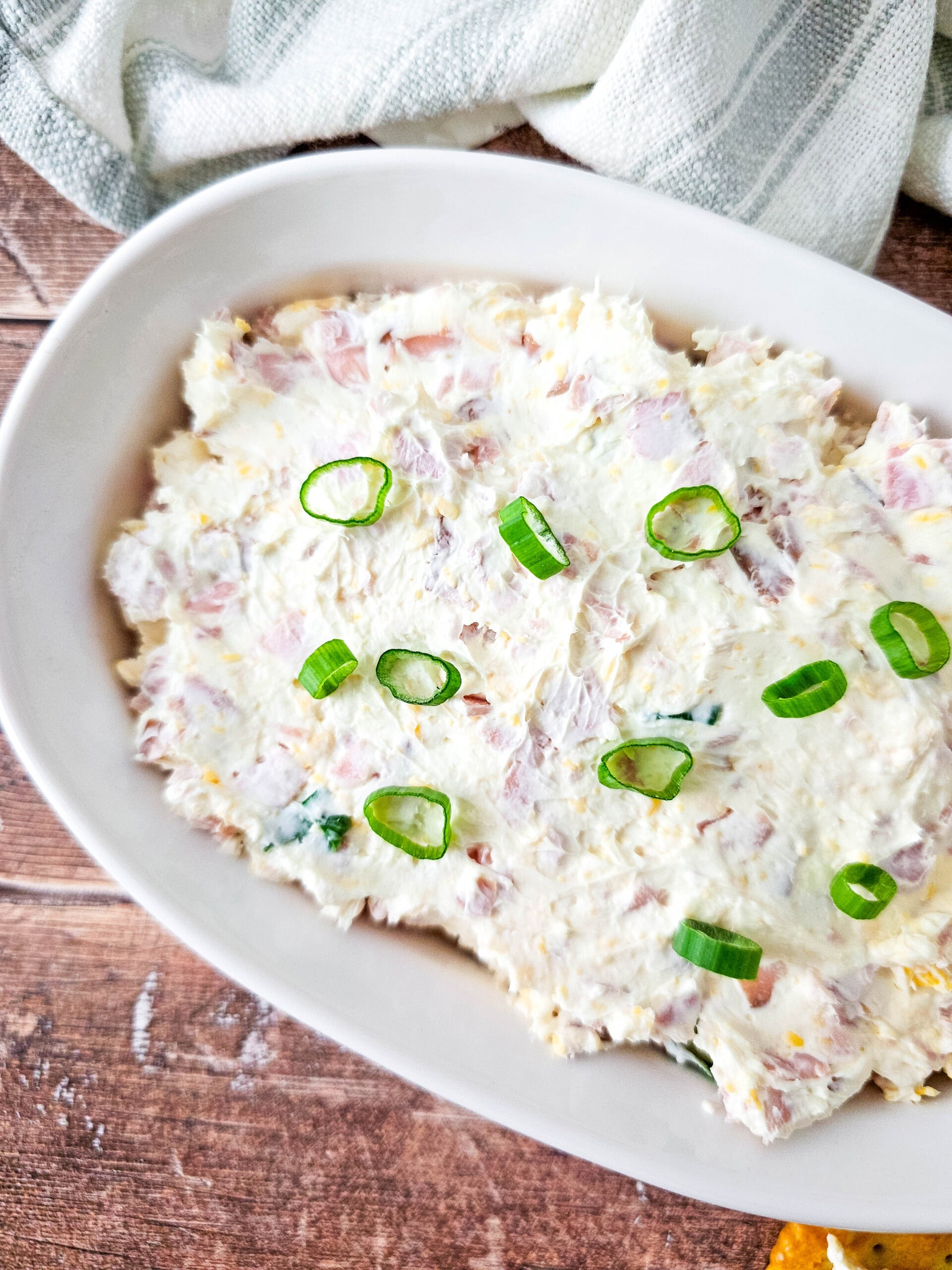An overhead of a white oval baking dish filled with creamy cheesy ham dip, topped with sliced green onions, with a soft kitchen towel in the background.