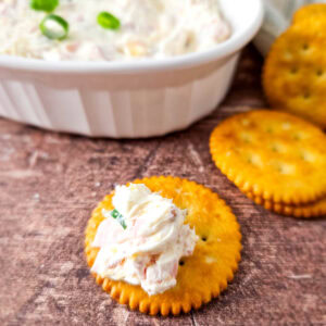 A cracker with a scoop of cheesy ham dip placed in front of a dish of dip and a small stack of crackers, all on a rustic wooden surface.