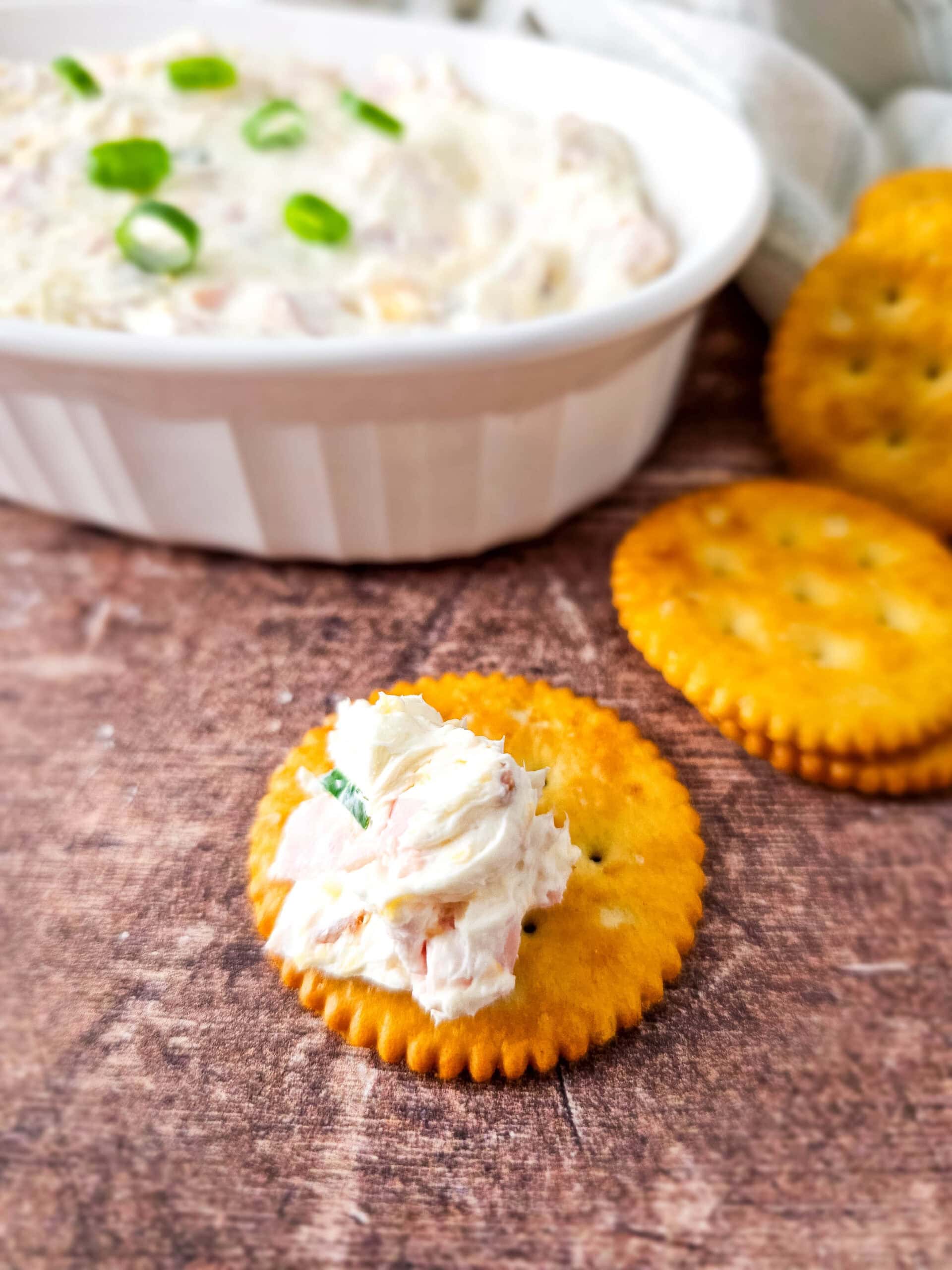 A cracker with a scoop of cheesy ham dip placed in front of a dish of dip and a small stack of crackers, all on a rustic wooden surface.