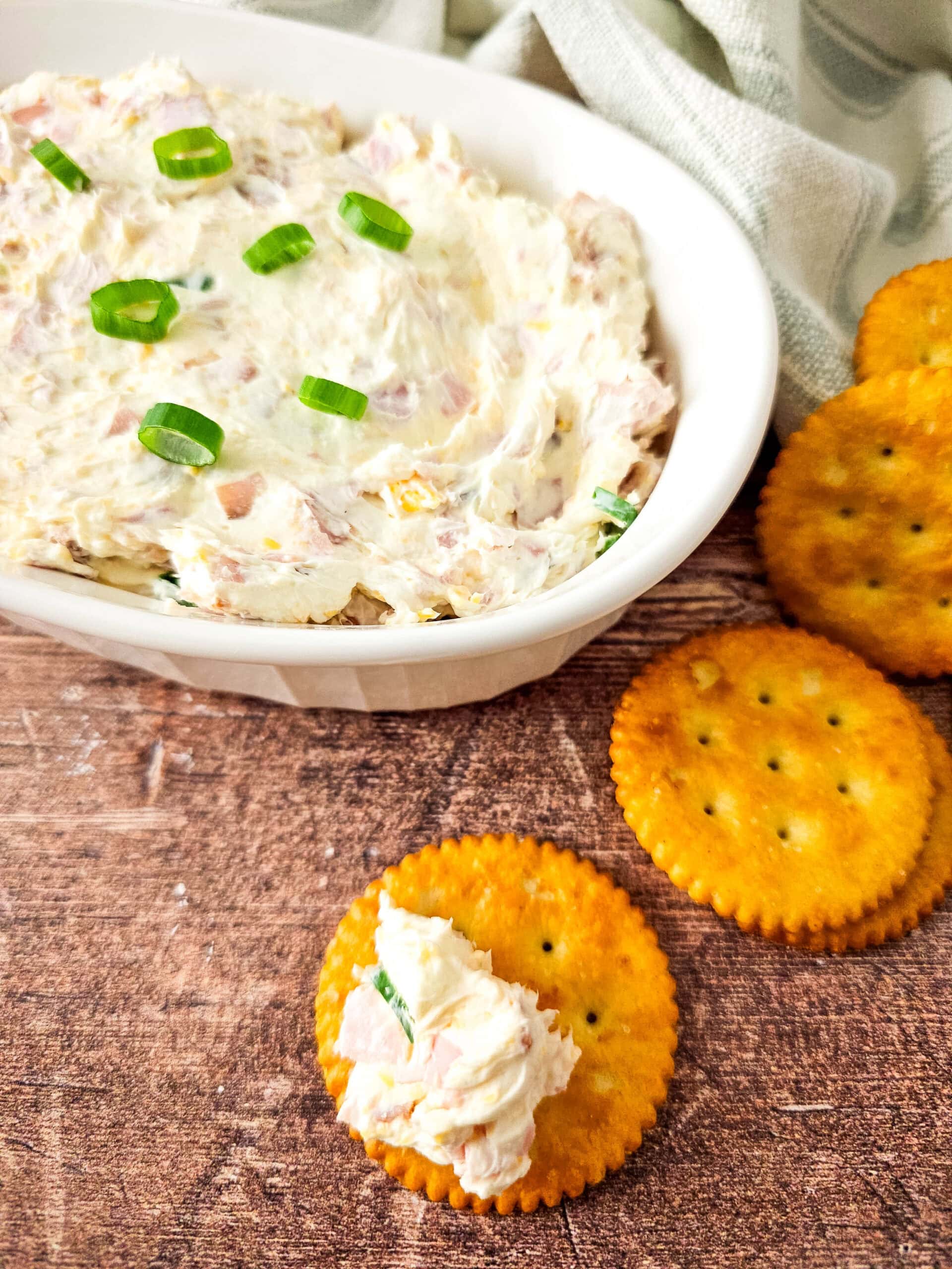 A dish of cheesy ham dip garnished with green onions, surrounded by round golden crackers, with one cracker topped with dip in the foreground.