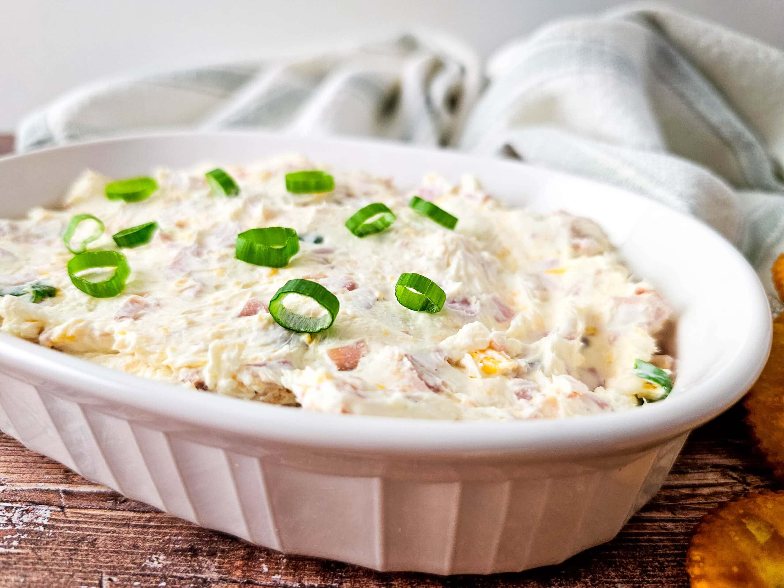 A white oval dish filled with creamy cheesy ham dip, topped with sliced green onions, sits on a rustic wooden surface next to several round crackers.