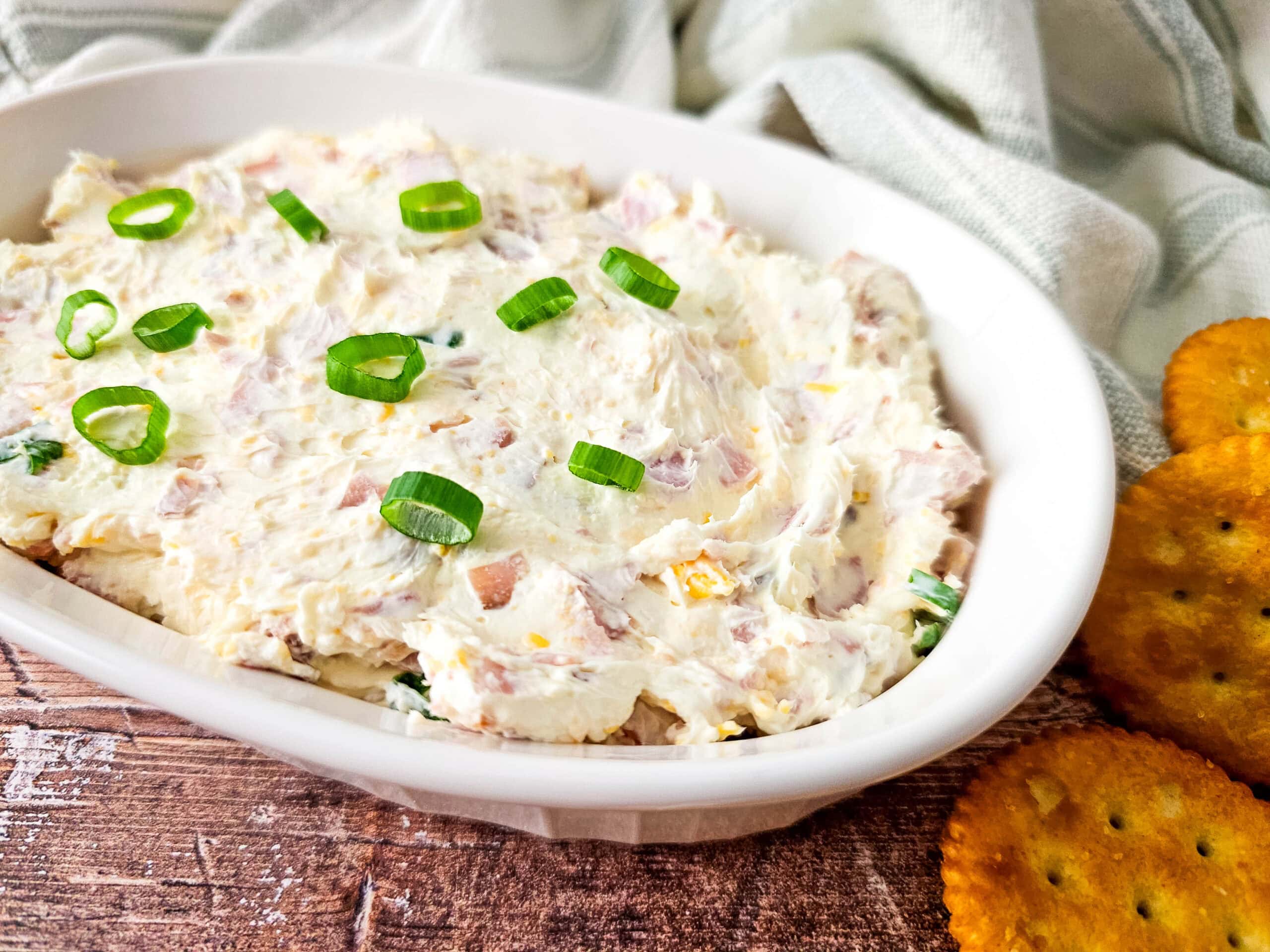 An overhead shot of a white oval baking dish filled with cheesy ham dip, garnished with sliced green onions, on a wooden table with a light striped kitchen towel in the background.