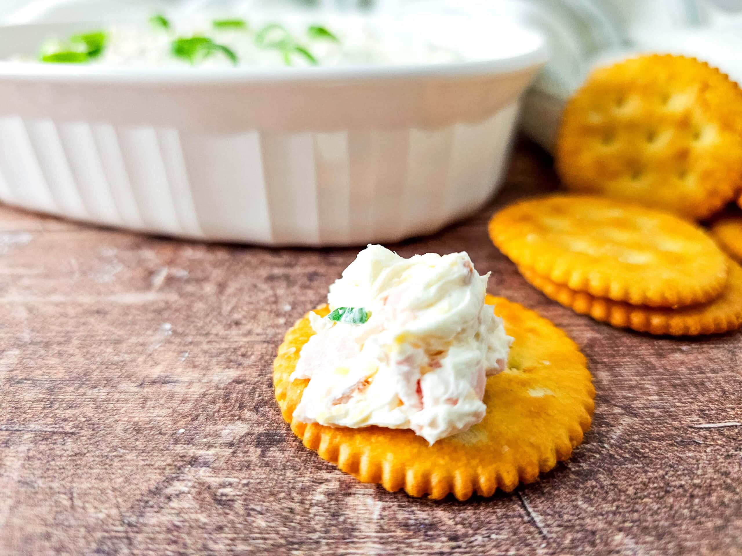 A white oval dish filled with creamy cheesy ham dip, topped with sliced green onions, sits on a rustic wooden surface next to several round crackers.
