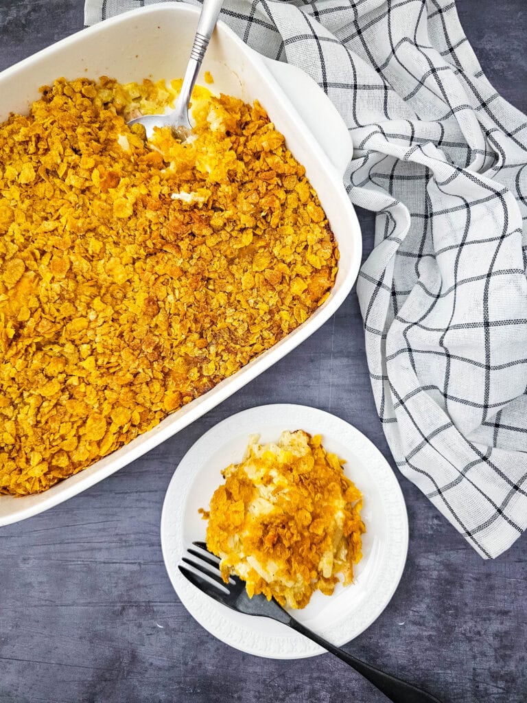 A close-up view of a hand holding a spoon scooping into a casserole dish of cheesy party potatoes with a crunchy golden topping.