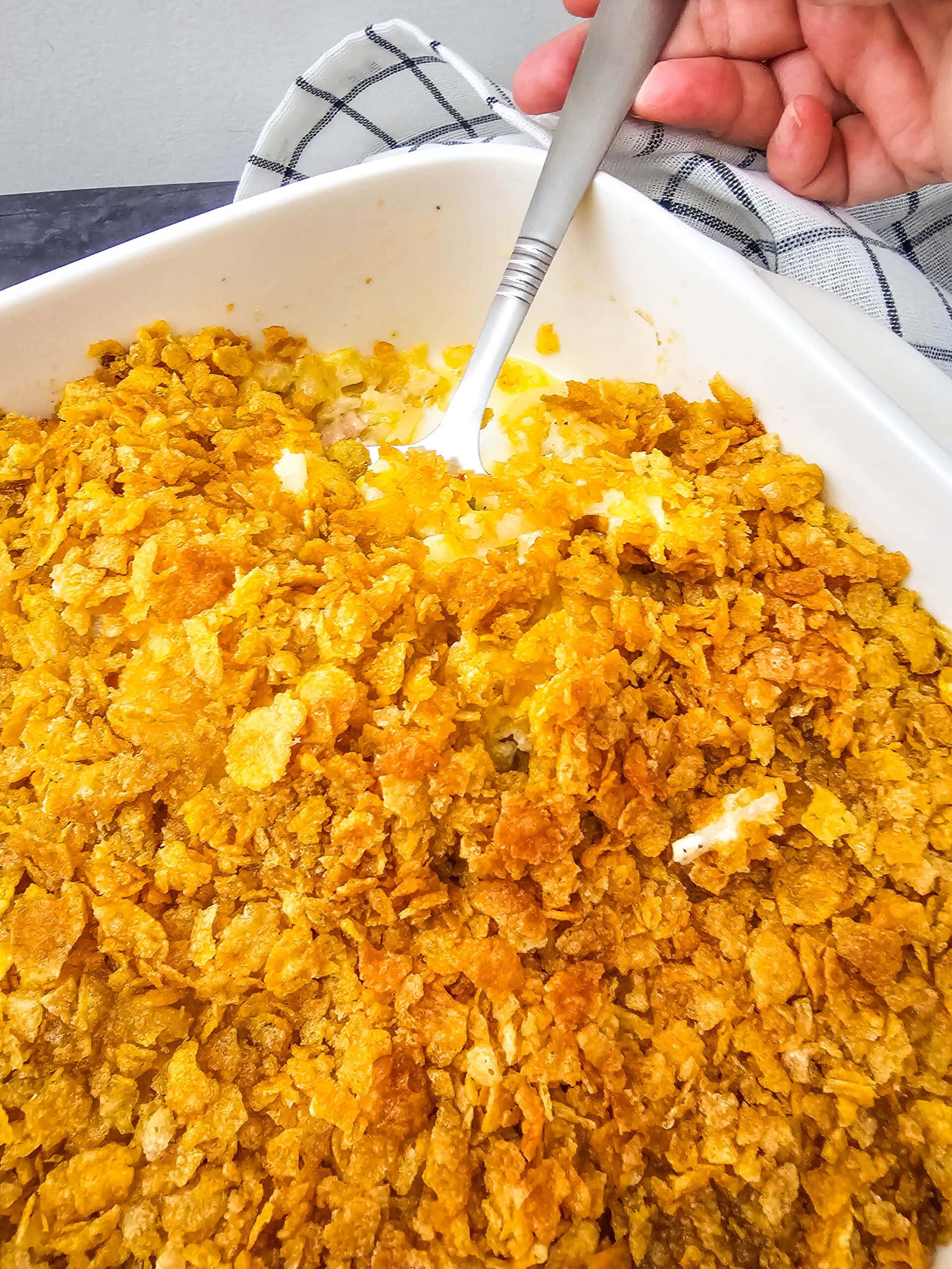 A close-up view of a hand holding a spoon scooping into a casserole dish of cheesy party potatoes with a crunchy golden topping.