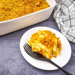 Close-up of a plated portion of cheesy party potatoes next to a white casserole dish, topped with a crispy cornflake crust.
