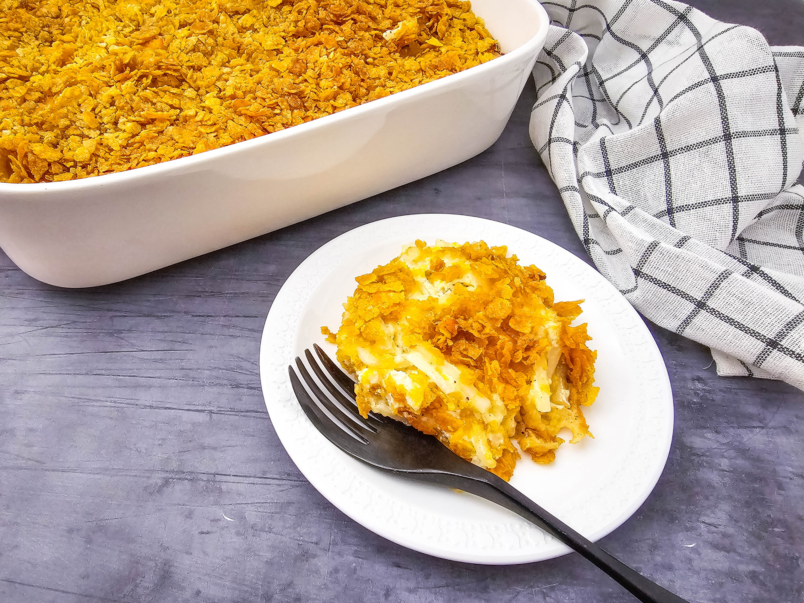 Close-up of a plated portion of cheesy party potatoes next to a white casserole dish, topped with a crispy cornflake crust.