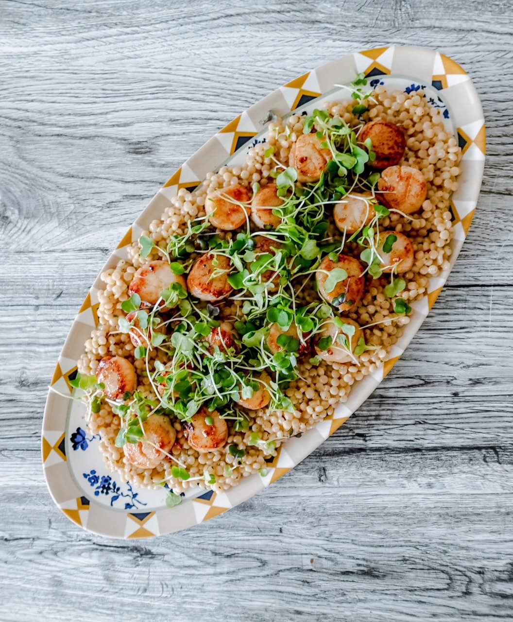 Overhead view of a long patterned platter filled with couscous, seared scallops, and vibrant microgreens on a rustic wooden table.