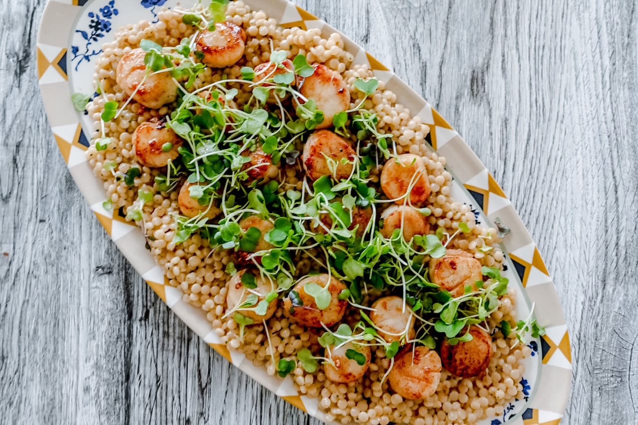 Angled shot of scallops arranged over couscous with microgreens scattered on top, capturing a beautifully styled seafood dish.