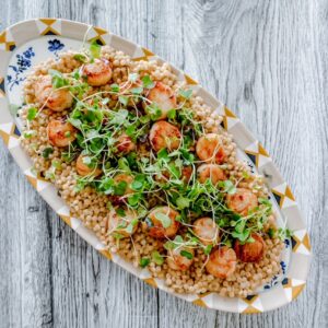 Wide top-down shot of a full serving platter featuring pearl couscous, seared scallops, and green garnish for a fresh, inviting presentation.