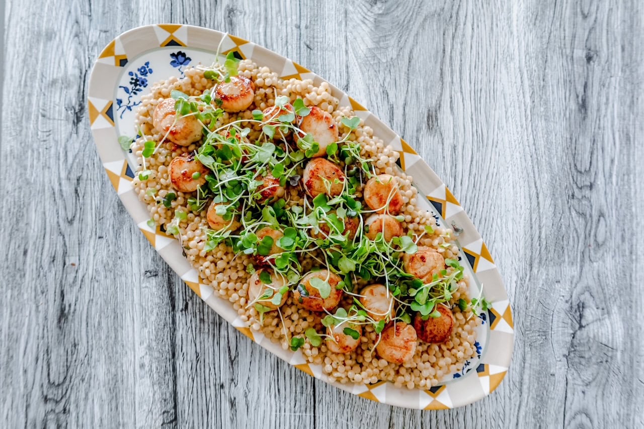 Wide top-down shot of a full serving platter featuring pearl couscous, seared scallops, and green garnish for a fresh, inviting presentation.