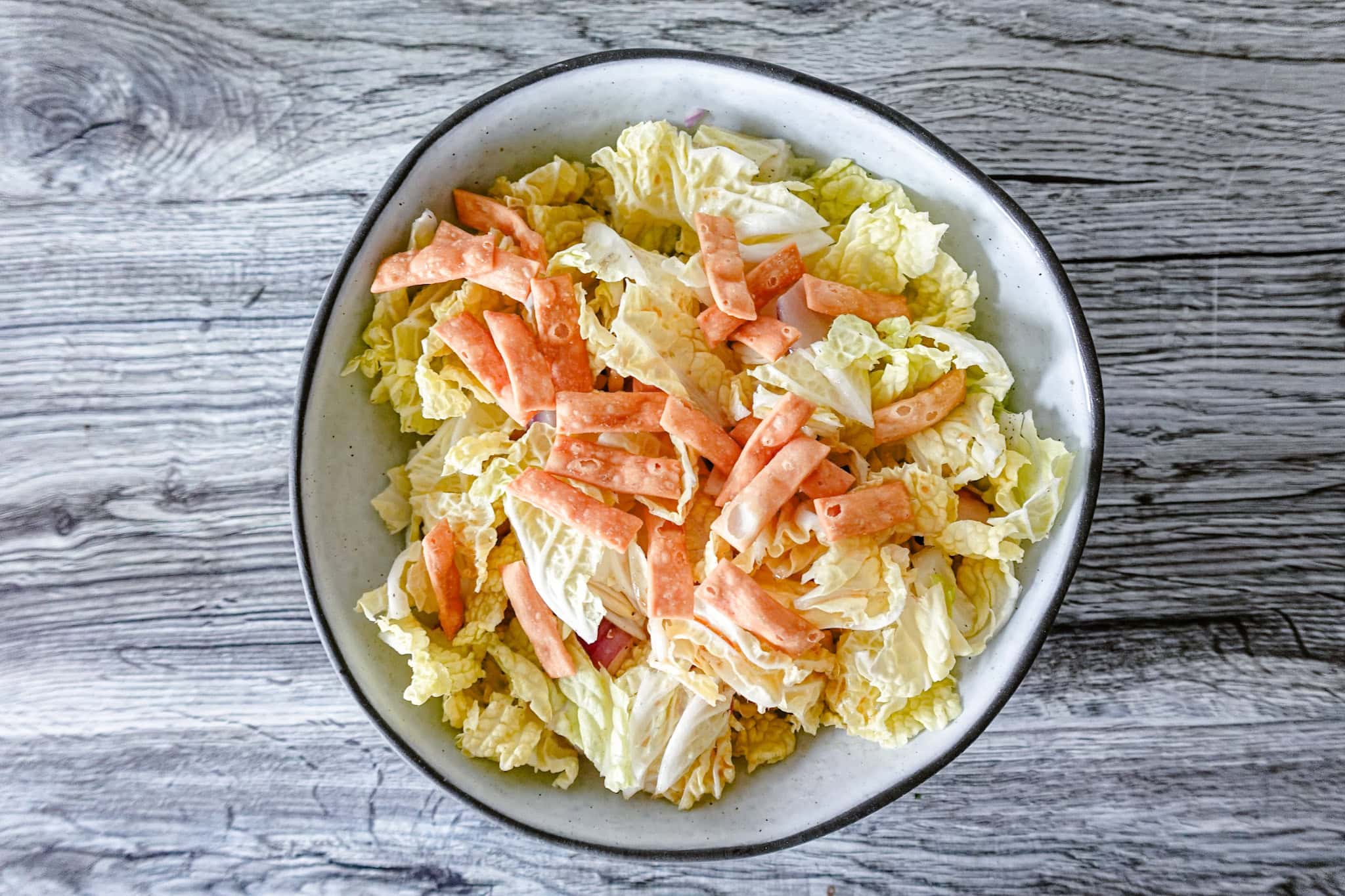 A top-down view of napa cabbage salad in a bowl, showing napa cabbage, crunchy wonton strips, and red onion on a wooden background.