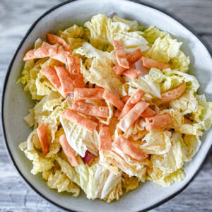 An overhead view of a white bowl filled with napa cabbage salad, garnished with crispy wonton strips and red onion slices, set on a gray wooden surface.