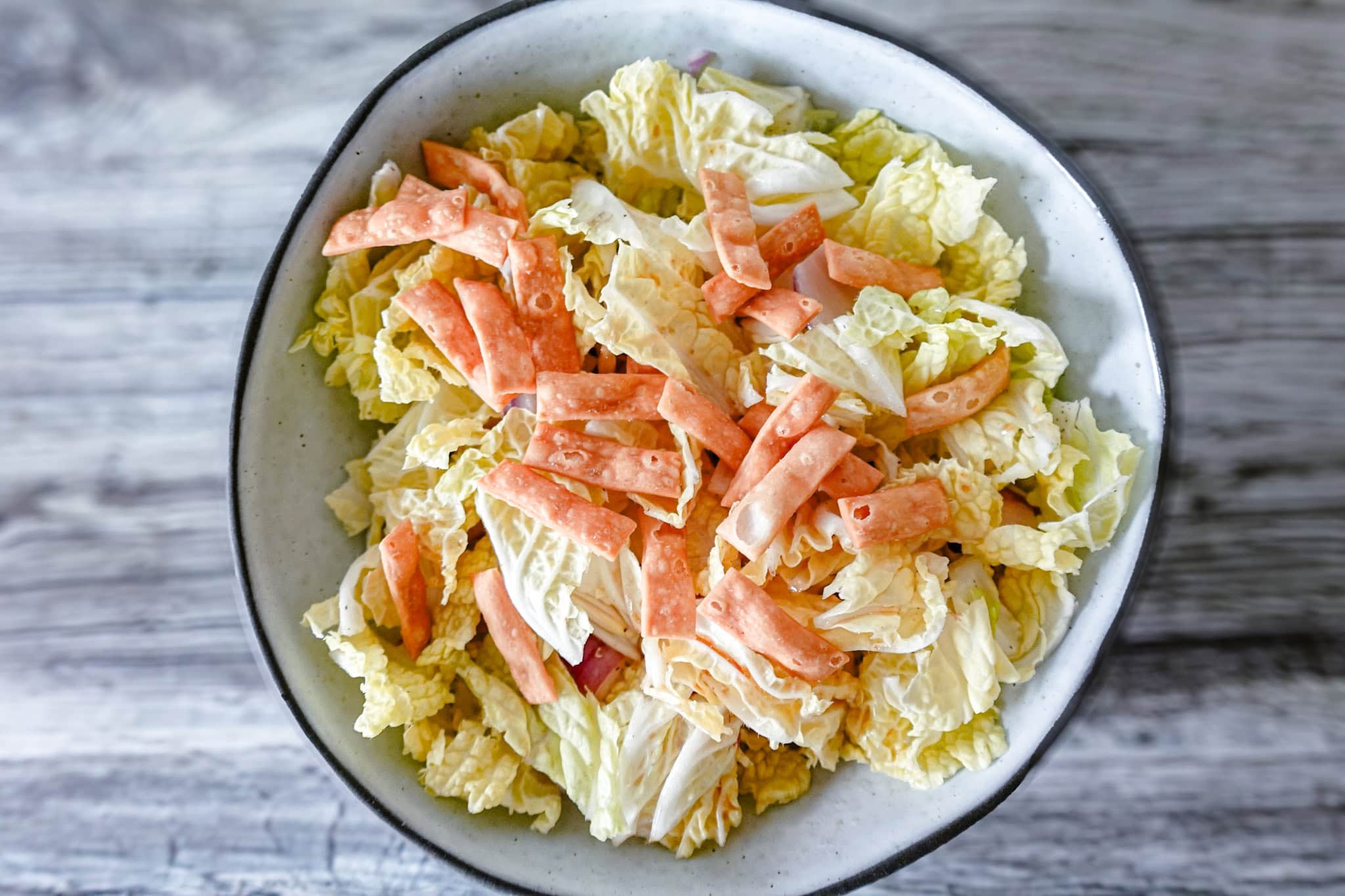 An overhead view of a white bowl filled with napa cabbage salad, garnished with crispy wonton strips and red onion slices, set on a gray wooden surface.