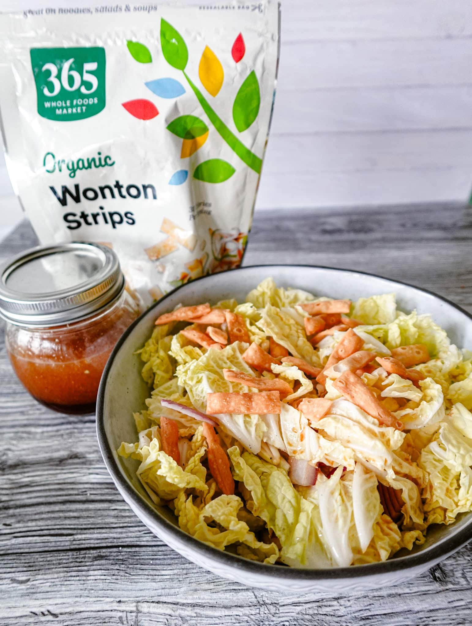 A bowl of napa cabbage salad sits on a table, flanked by a jar of homemade dressing and a bag of organic wonton strips in the background, emphasizing the salad's fresh and crunchy toppings.