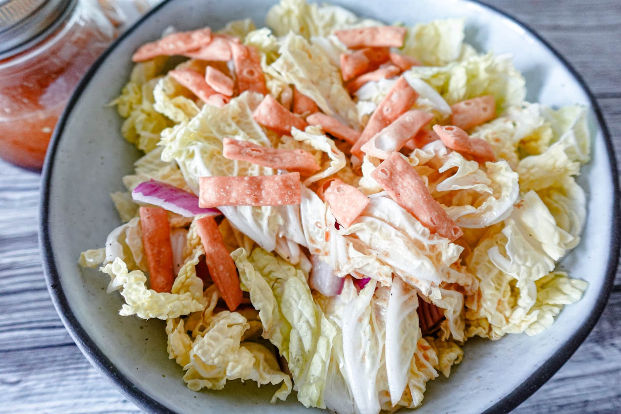 A close-up of a bowl filled with fresh napa cabbage salad, topped with crunchy wonton strips and thinly sliced red onions, with a jar of dressing in the background.