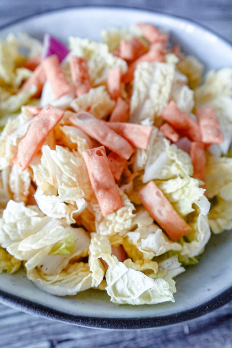 A vertical shot of napa cabbage salad in a white bowl, highlighting crisp cabbage leaves, carrot strips, red onion, and crunchy wonton strips.